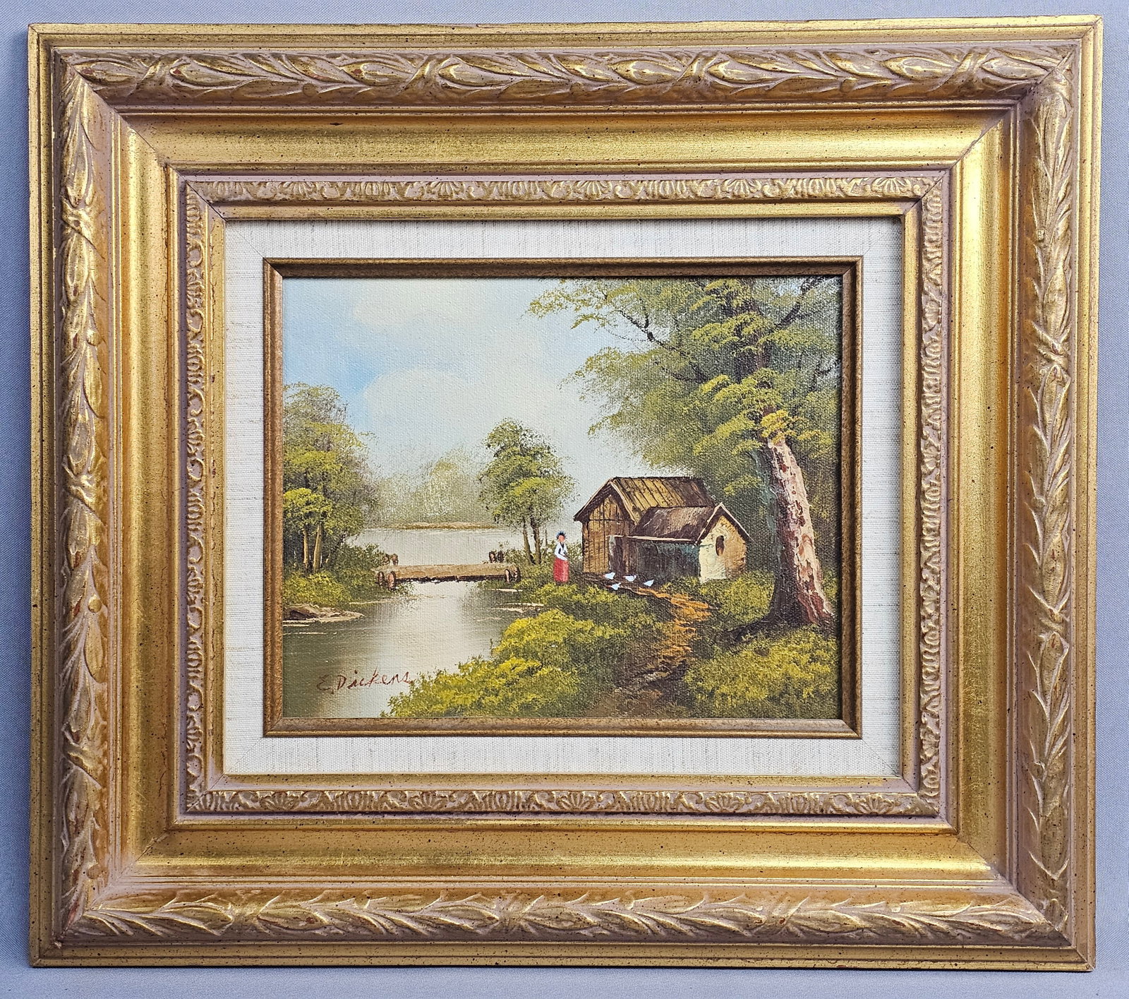 E. Dickens River Landscape with Cottage Oil on Canvas: This original oil painting on canvas by E. Dickens depicts a rustic river landscape featuring a timber cottage and a figure dressed in a red skirt standing at the water's edge. The signature E.