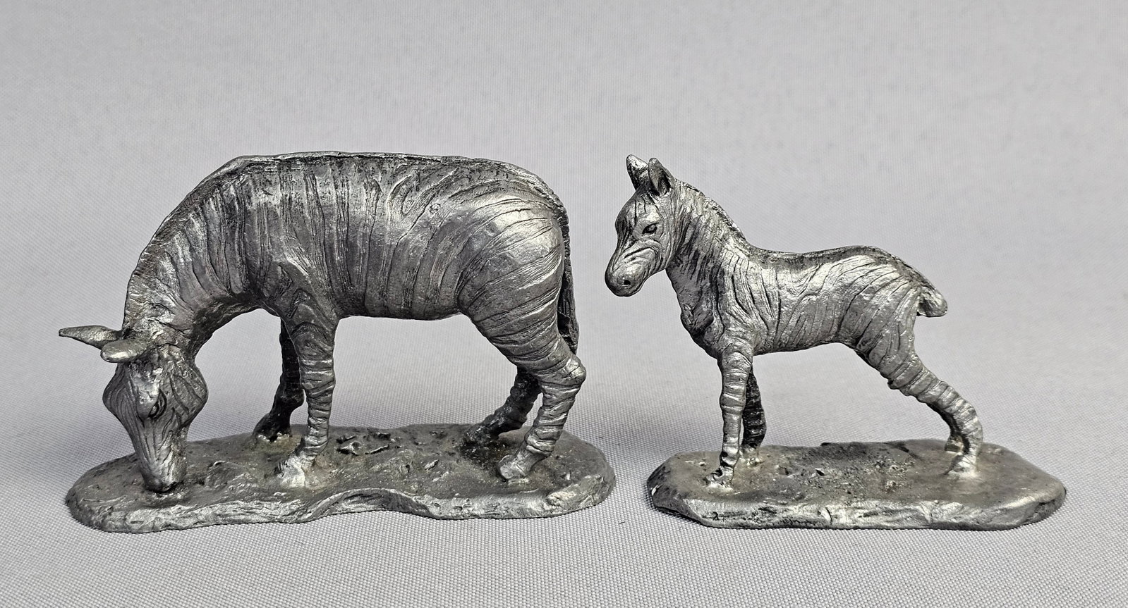 A Pair of Ricker-Bartlett Pewter Zebra Figurines: This lot features two cast pewter figurines from Ricker-Bartlett Pewter depicting a zebra family. The pair includes an adult zebra in a grazing stance and a standing foal, both mounted on integrated