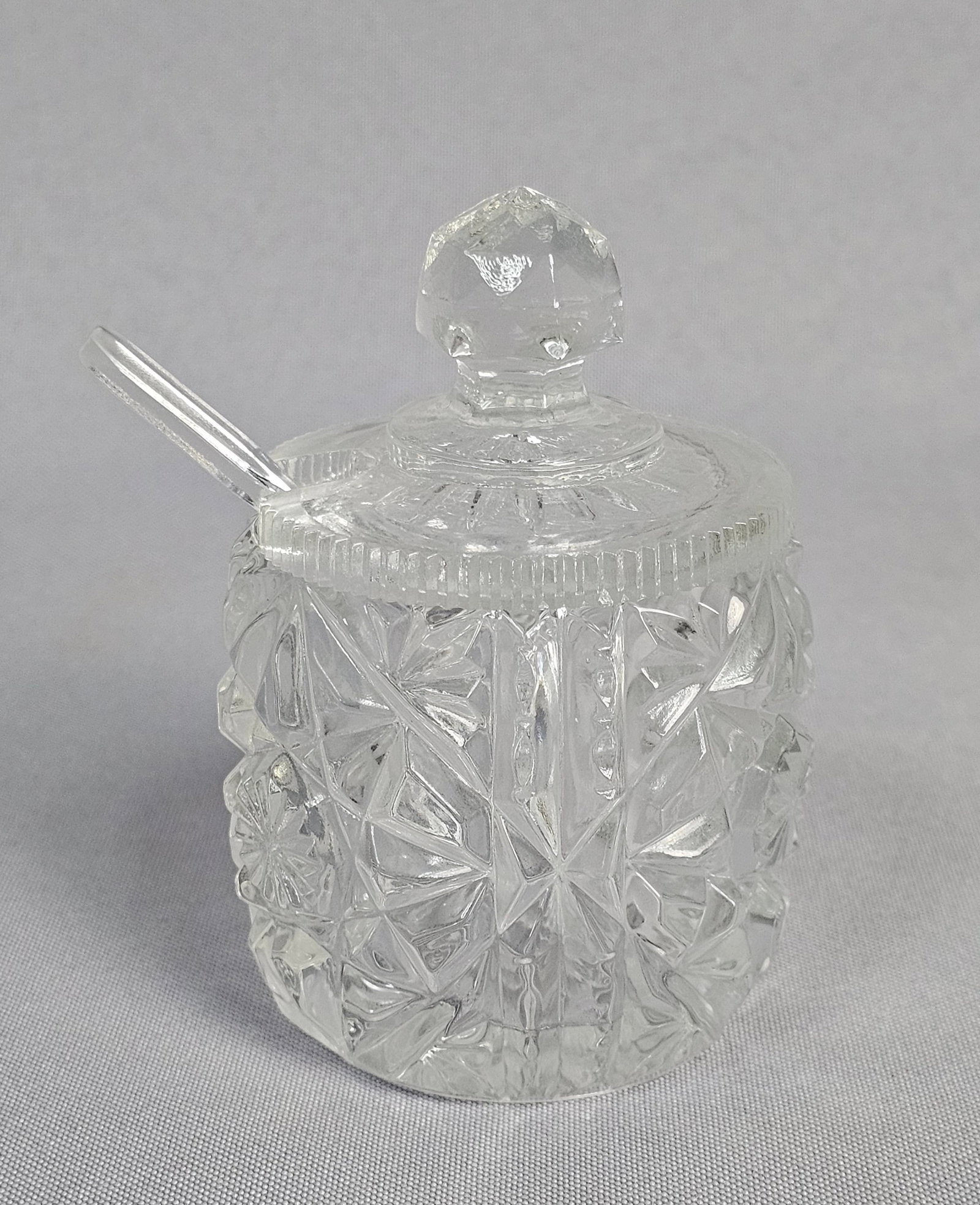 Pressed Glass Lidded Condiment Jar and Spoon: A clear pressed glass lidded condiment jar and coordinating spoon. The set is attributed to Anchor Hocking based on visual pattern identification. The cylindrical body is decorated with a repeating mo