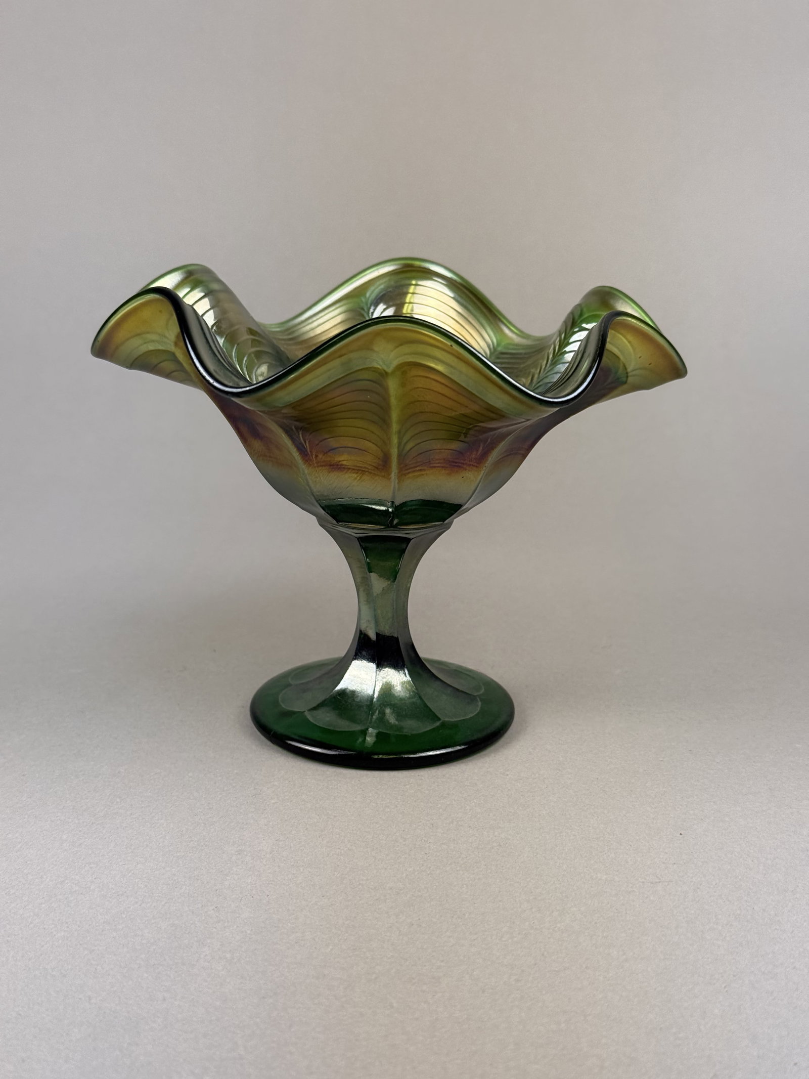 Antique Green Carnival Glass Ruffled Pedestal Compote: This piece is a pressed green glass compote featuring a vibrant iridescent carnival finish. The interior of the bowl displays the Peacock Tail pattern. The upper surface of the foot is decorated with 