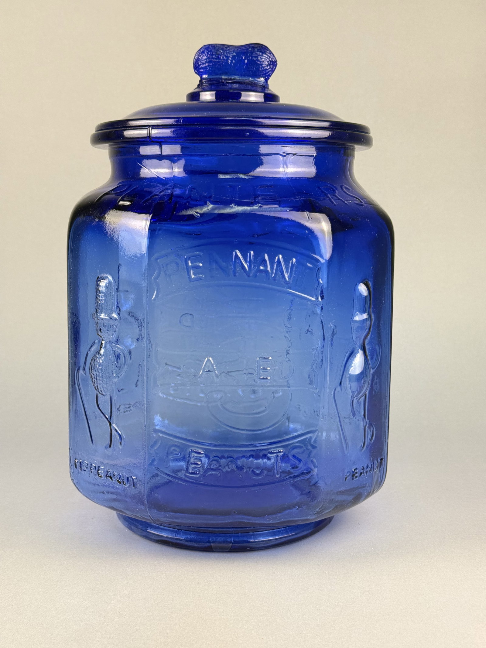 Large Cobalt Blue Glass Planters Peanuts Pennant 5 Cent Display Jar: This counter display jar for Planters Peanuts is made of deep cobalt blue molded glass. Two side panels are decorated with embossed figures of the Mr. Peanut mascot, complete with a top hat, monocle,