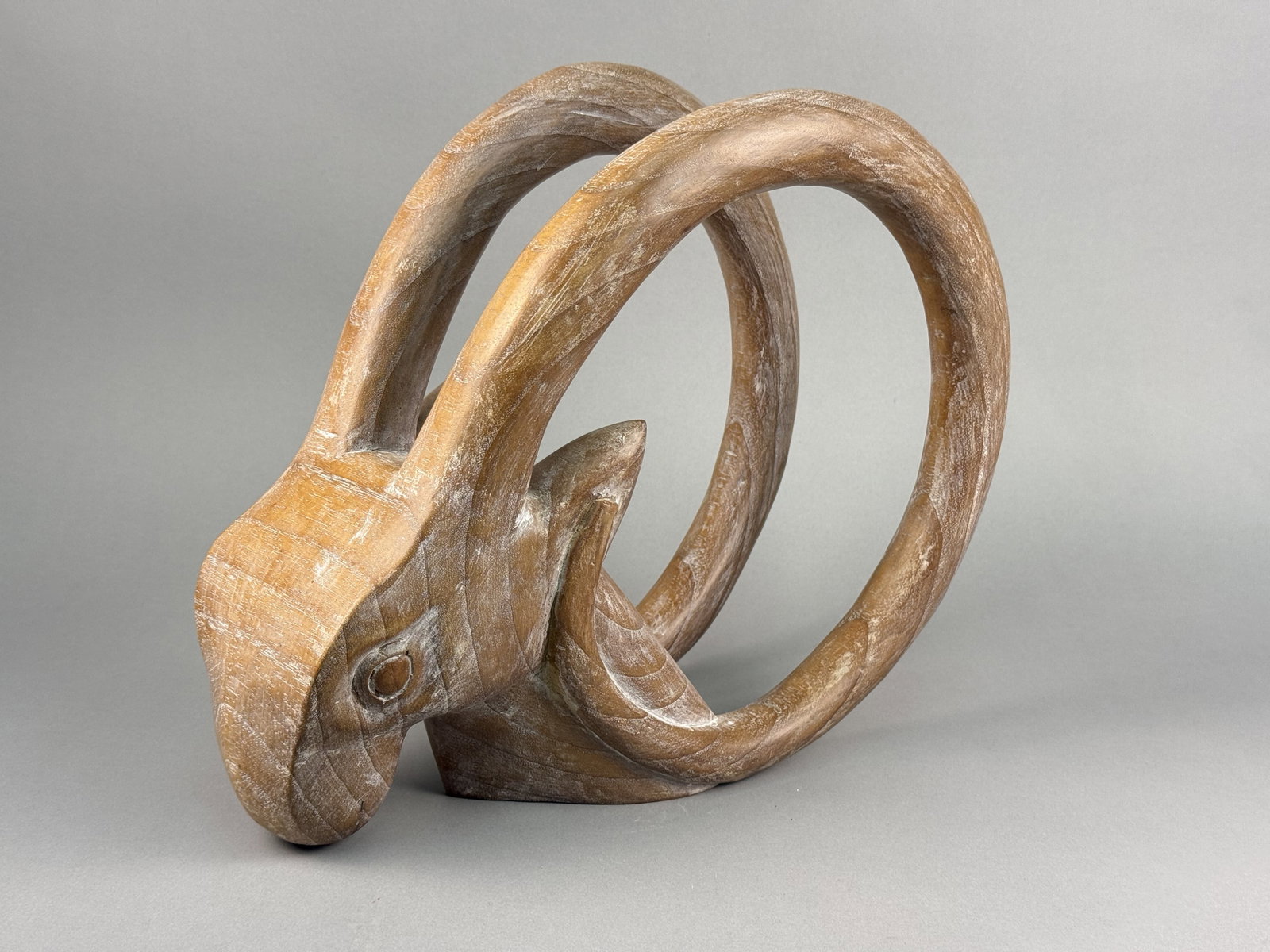 Modernist Stylized Carved Wood Ibex Head Sculpture: This modernist sculpture is hand-carved from wood, featuring a highly stylized depiction of an ibex or mountain goat head. The sculpture stands on a flat, integral base. No signatures, maker marks, or