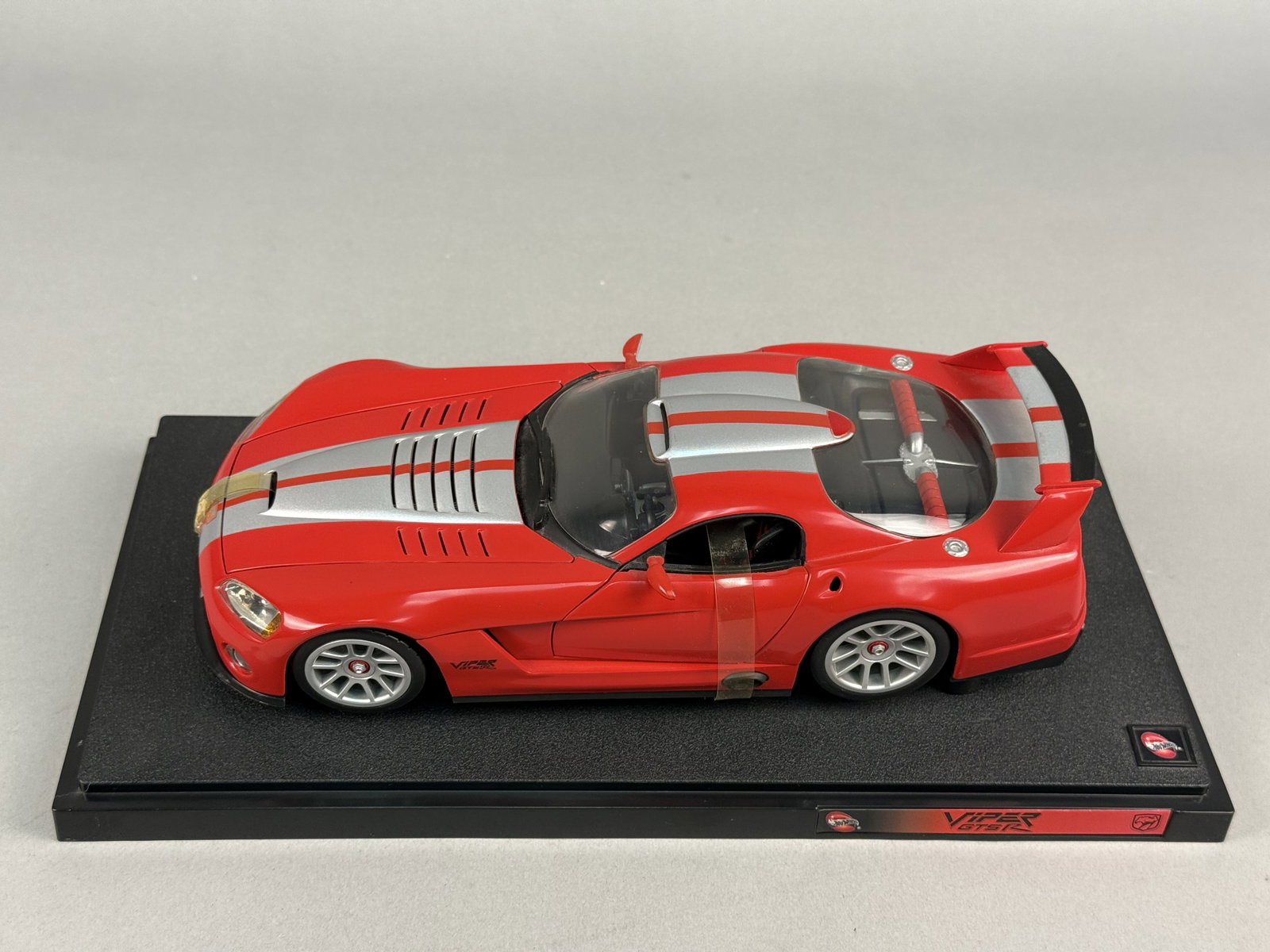 1:18 Scale 100% Hot Wheels Dodge Viper GTS-R Die-Cast Model Car: This 1:18 scale die-cast model car by 100% Hot Wheels depicts a Dodge Viper GTS-R. The model is mounted on a black textured plastic display base. Printed labels on the front edge of the base include t