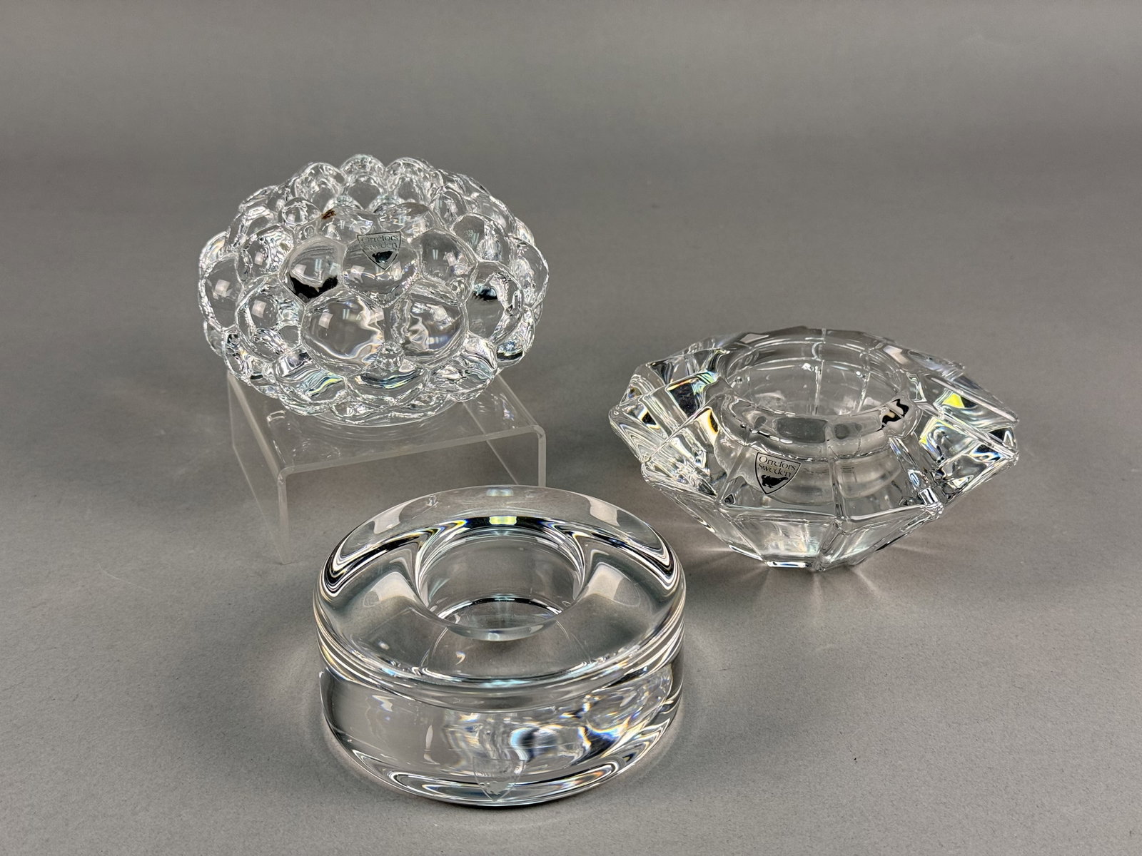 Trio of Orrefors Crystal Candle Holders Inc Raspberry Design: A trio of clear crystal glass candle holders produced by the Swedish glassworks Orrefors. The collection features three distinct designs: a globular Raspberry votive designed by Anne Nilsson; a low, c