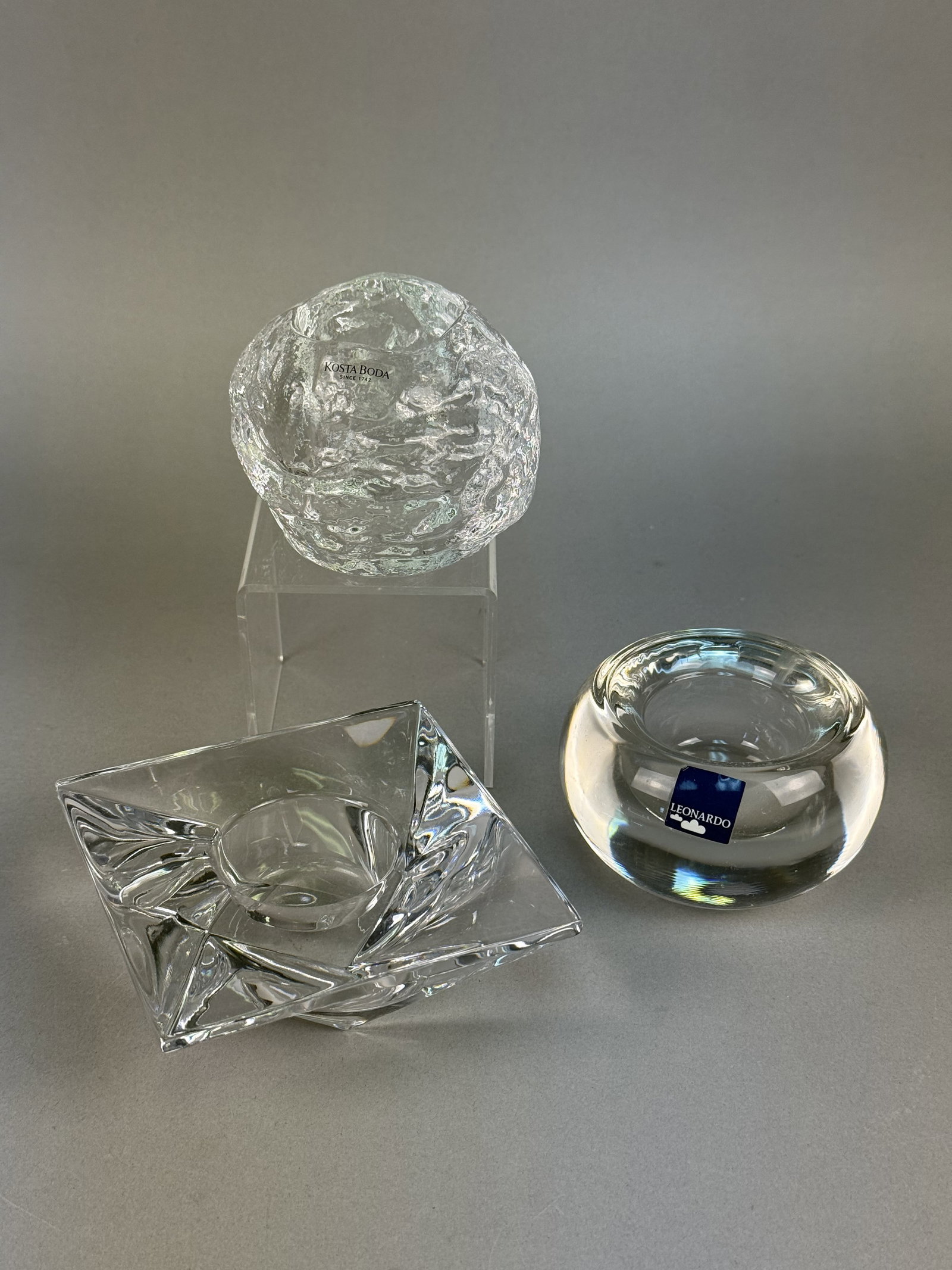 Art Glass Votive Candle Holders Inc Kosta Boda, Miller: This lot features three clear art glass votive or tea light candle holders from notable international glassmakers. The group includes a heavy Swedish crystal holder by Kosta Boda with a deeply texture