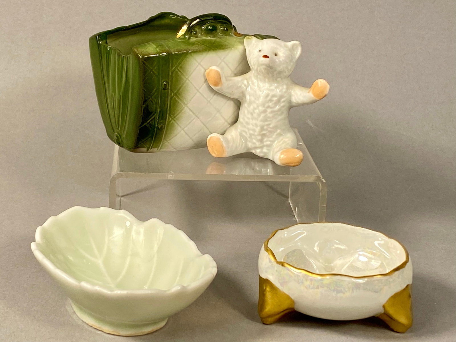 Three Miniature Porcelain Decorative Vessels: This collection features three miniature porcelain decorative pieces, including a vessel in the form of a suitcase with an attached bear, a footed iridescent bowl, and a leaf-shaped dish. The
