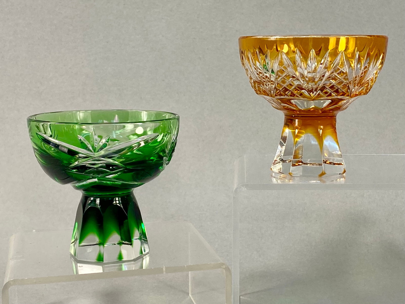 Pair of GEHA Cut-to-Clear Lead Crystal Footed Bowls: A pair of small, hand-cut lead crystal footed bowls produced by the German glass manufacturer GEHA. This pair consists of one emerald green and one amber-colored example, both featuring cased glass wi