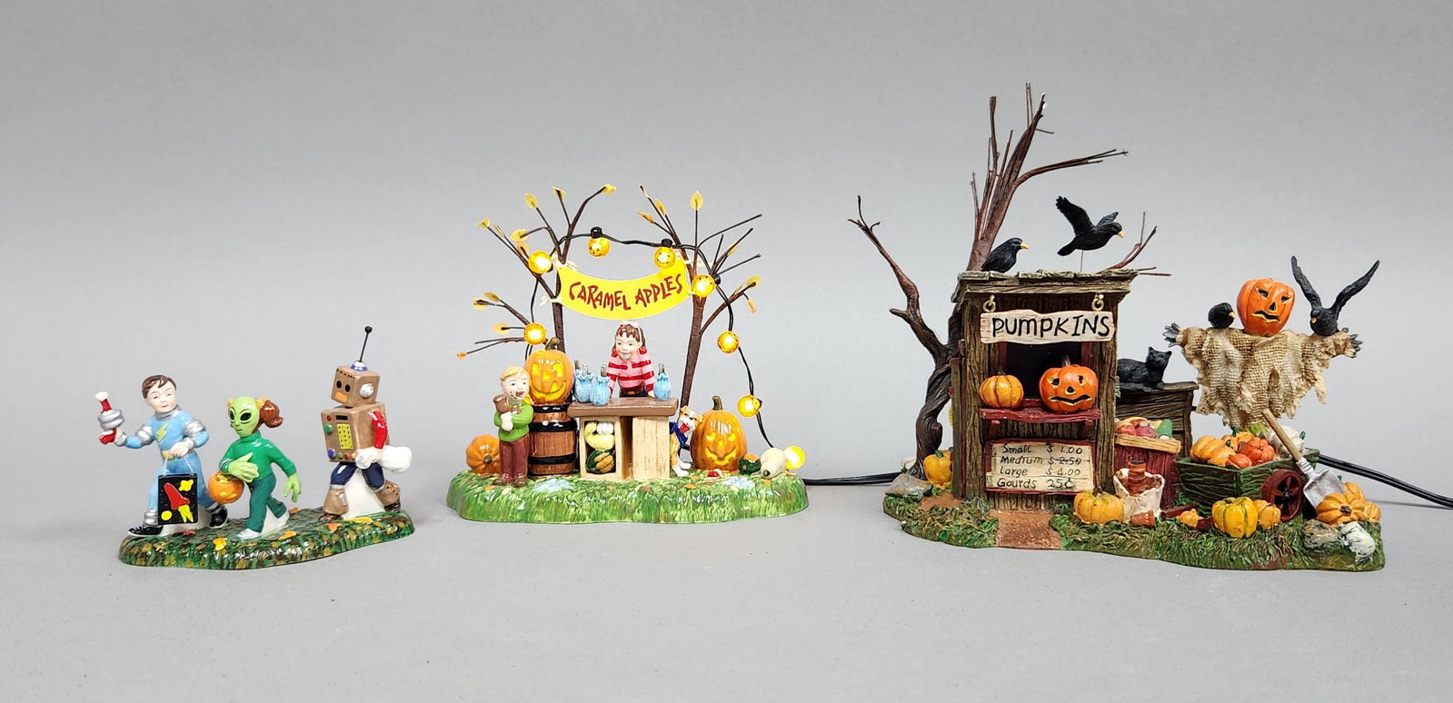 Dept 56 Halloween Village Accessories: This lot features a collection of three hand-painted ceramic figurines designed for seasonal village displays. Trick-or-treating trio height: 2.75 inches Caramel apple stand height: 4.75 inches