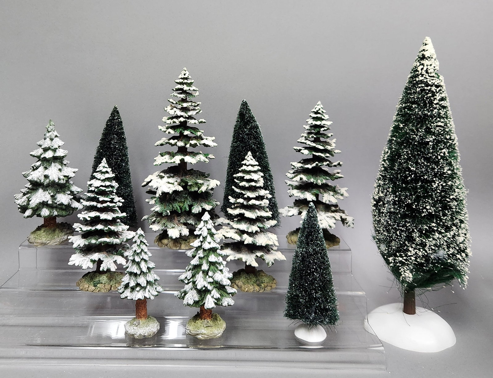 Dept 56 Pequot Pines & Bottle Brush Trees: A collection of various miniature decorative evergreen trees produced by Department 56. The assortment includes snow-dusted resin models from the Village Pequot Pines series and bottle-brush