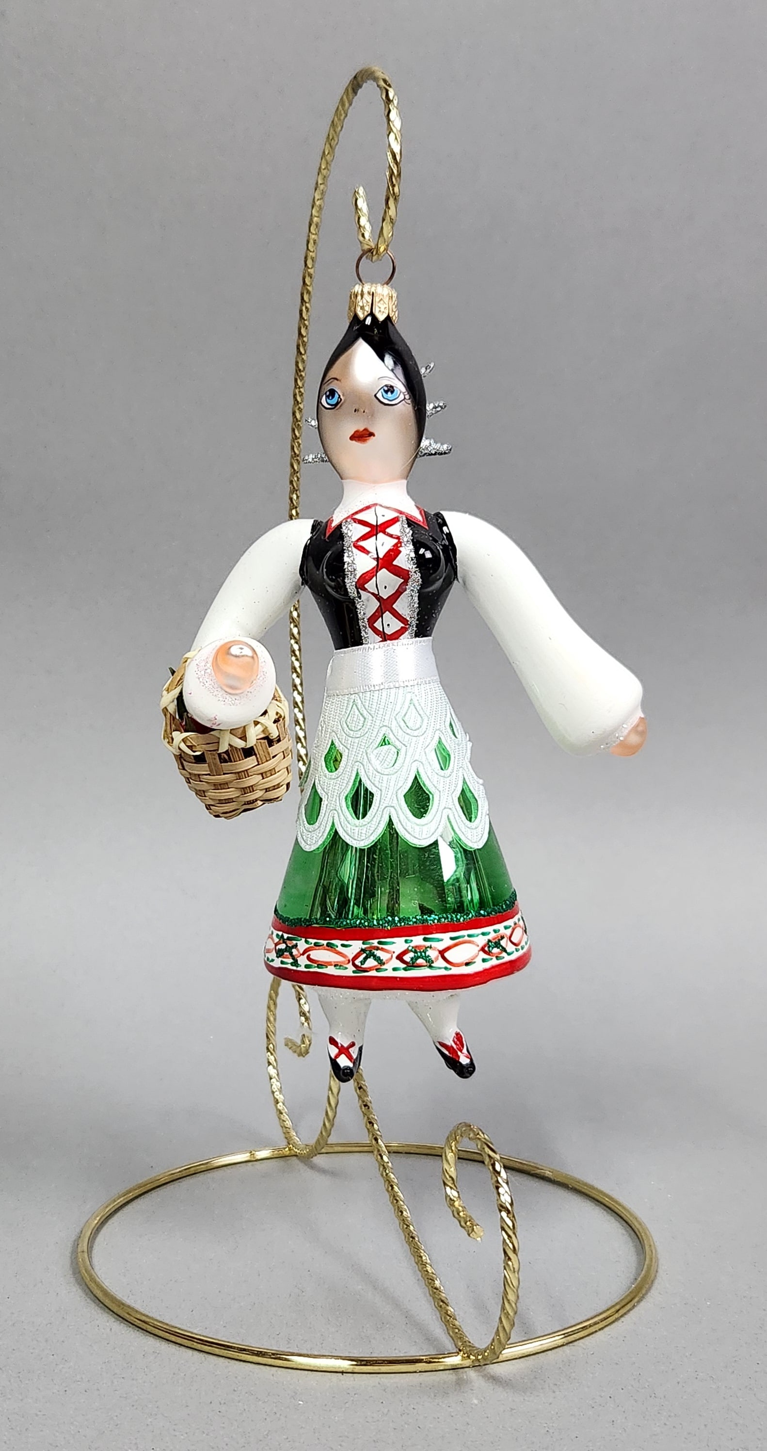 Glass European Folk Woman Ornament: Stand not included. A hand-blown glass Christmas ornament depicting a woman in traditional European folk dress with a green skirt and woven basket, measuring approximately 6.75 inches in height. Very