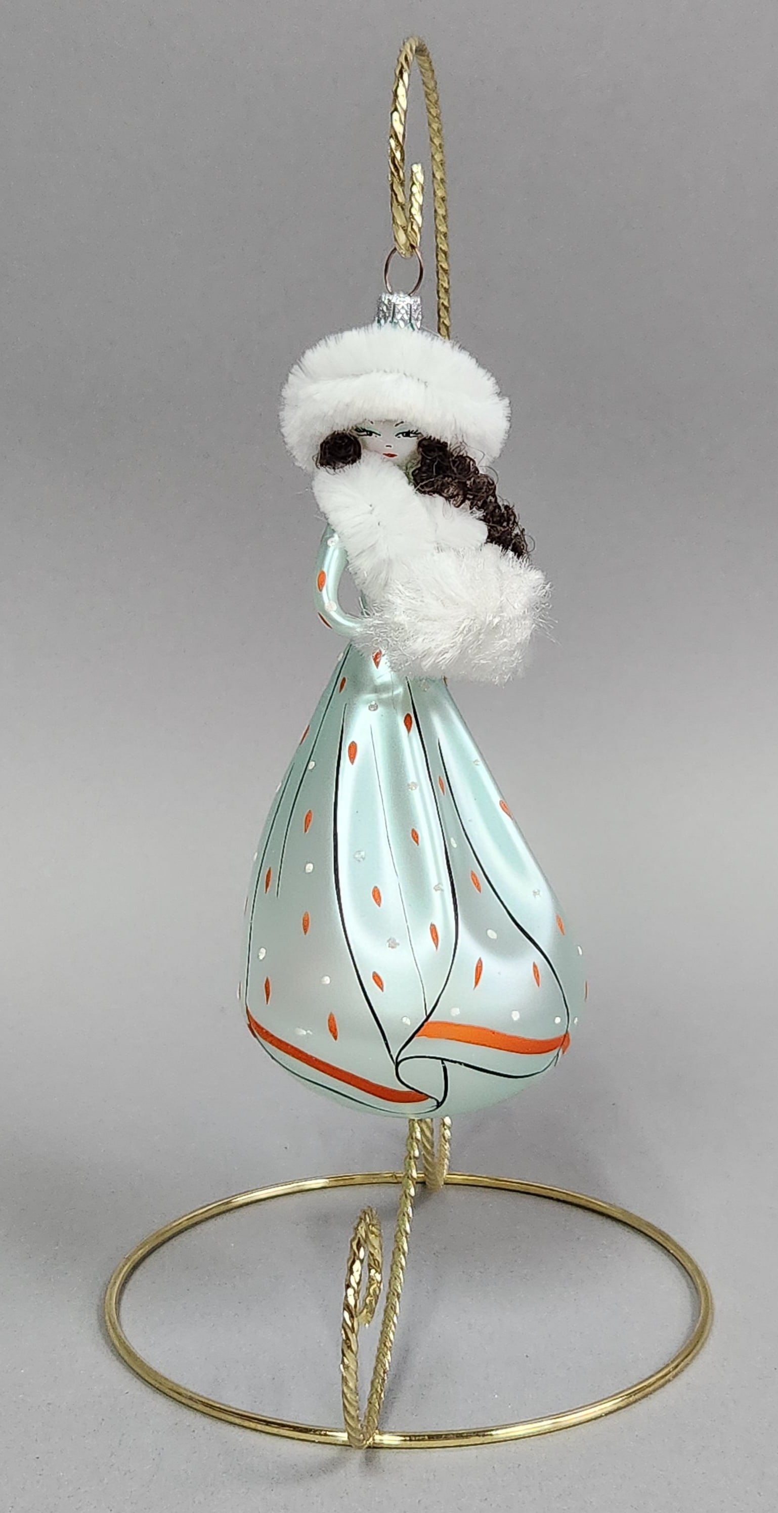 De Carlini Heiress Glass Ornament: Stand not included. This hand-blown glass Heiress Maris Wintrelle Christmas ornament is shaped as a stylized female figure in winter attire. MADE and ITALY. Length: approximately 6 inches. The