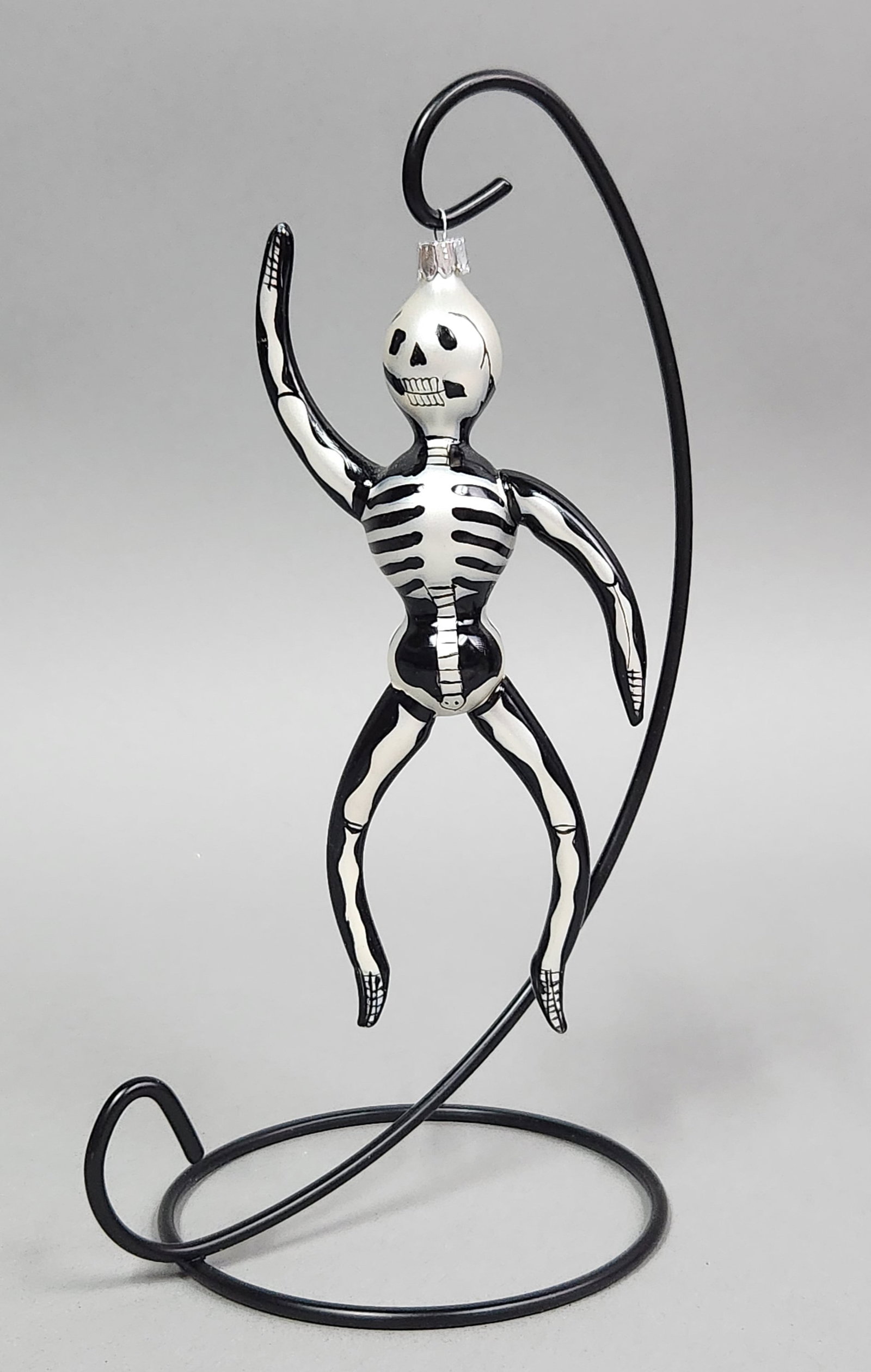 Glass Skeleton Ornament: Stand not included. A hand-painted black and white glass skeleton ornament measuring approximately 6 inches in length, accompanied by a black metal hanging stand. Very good condition.