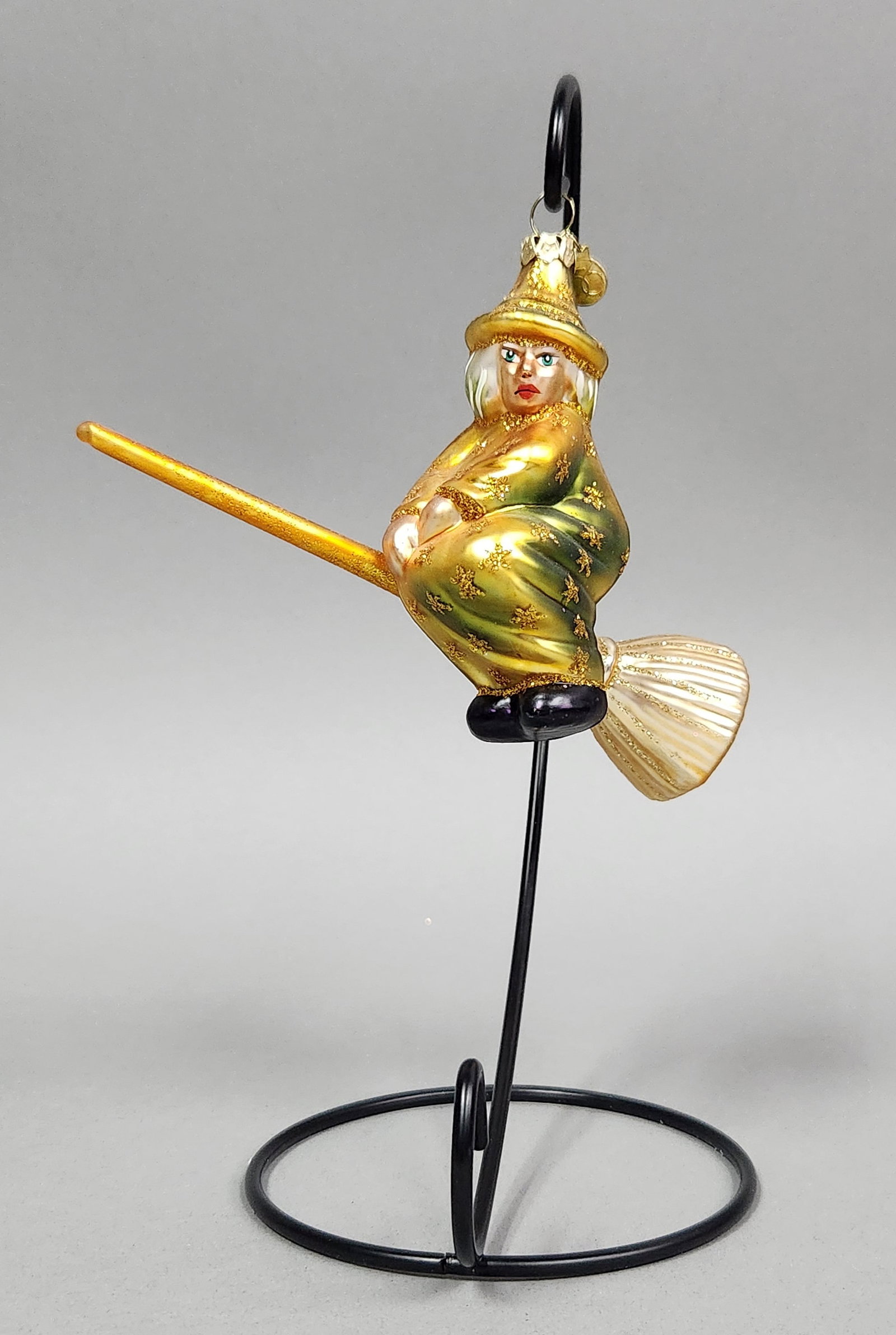 Radko Glass Witch Ornament: Stand not included. A Christopher Radko blown glass witch ornament in gold and green with glitter accents and a signature charm, measuring approximately 7.5 inches in length. Very good condition.