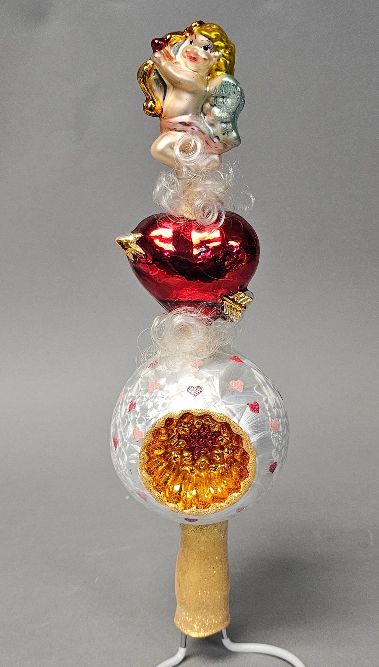 Radko Glass Cupid Valentines Tree Topper Finial: A three-tier vertical glass tree topper featuring a figural finial, a central heart motif, and a large reflector base orb. The ornament is constructed from hand-blown glass with a silvered interior fo
