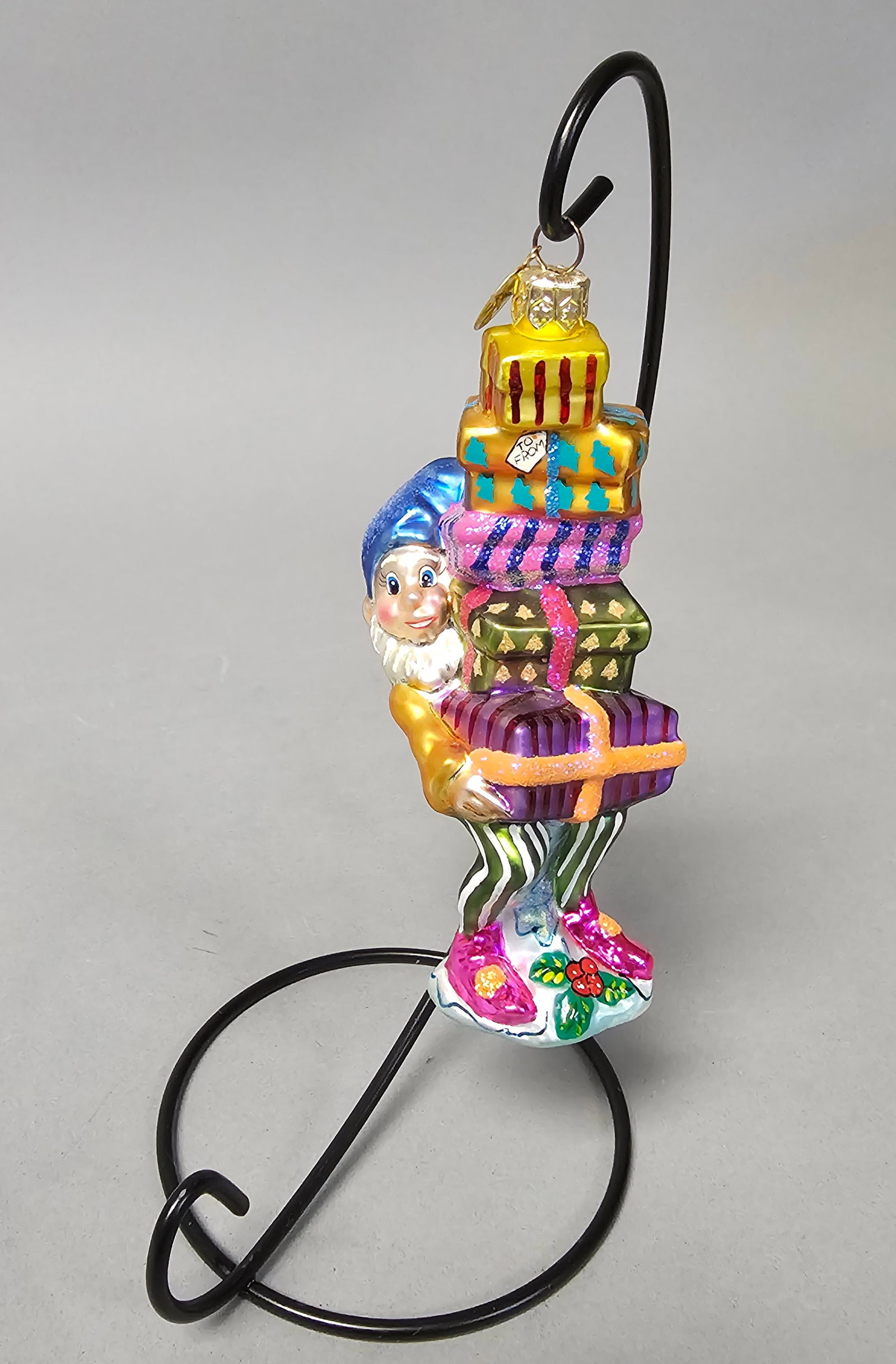 Radko Balancing Act Elf & Gifts Glass Ornament: *STAND NOT INCLUDED* A Christopher Radko Balancing Act handcrafted blown glass Christmas ornament. The figure depicts a whimsical elf with a white beard and shimmering blue hat carrying a precarious v