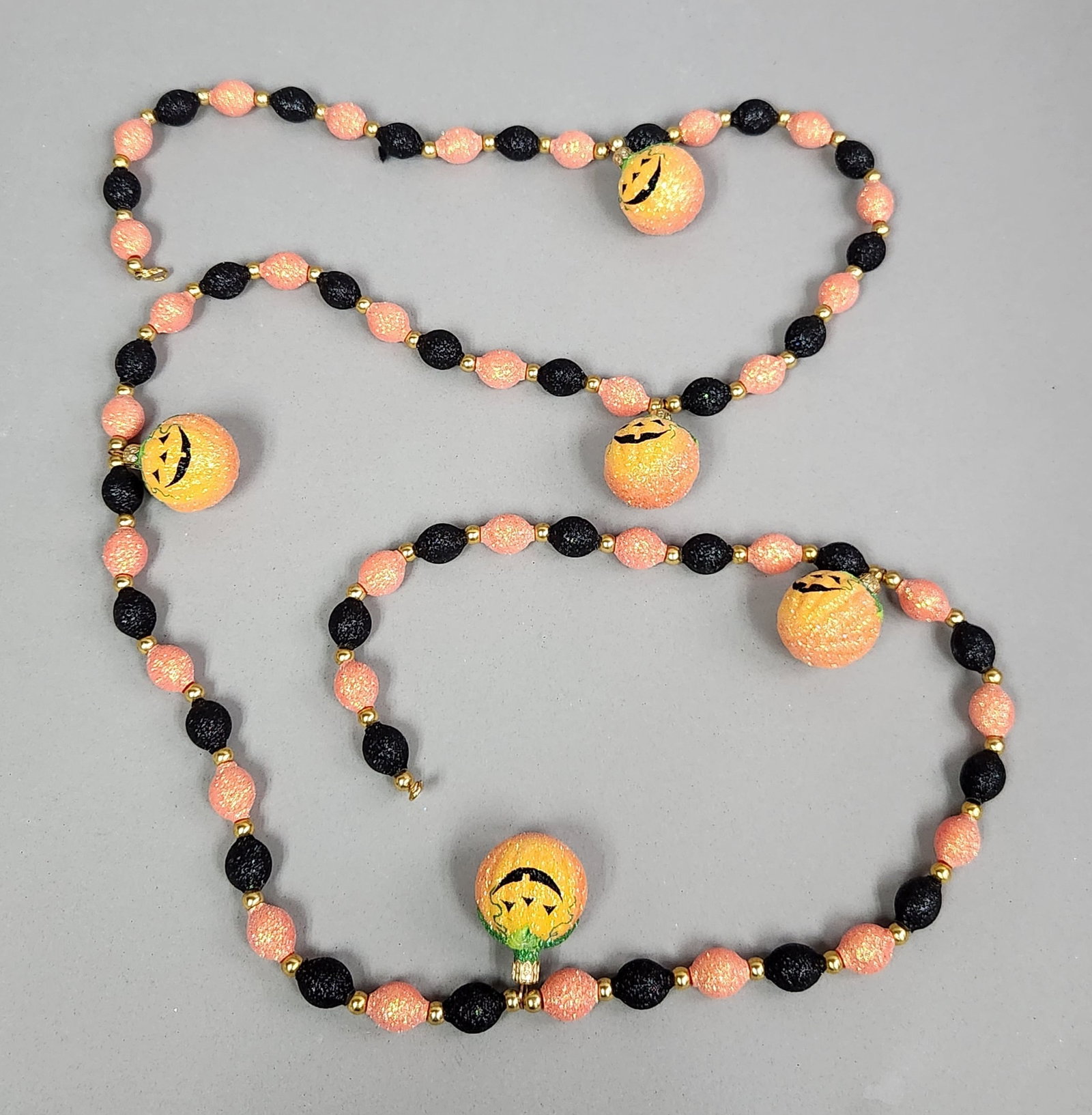 Halloween Jack-o'-Lantern Garland: A Halloween-themed decorative garland featuring alternating orange and black glitter-coated oval beads. The piece is finished with a metal hook-and-eye style clasp. Length: 75 inches. The garland is