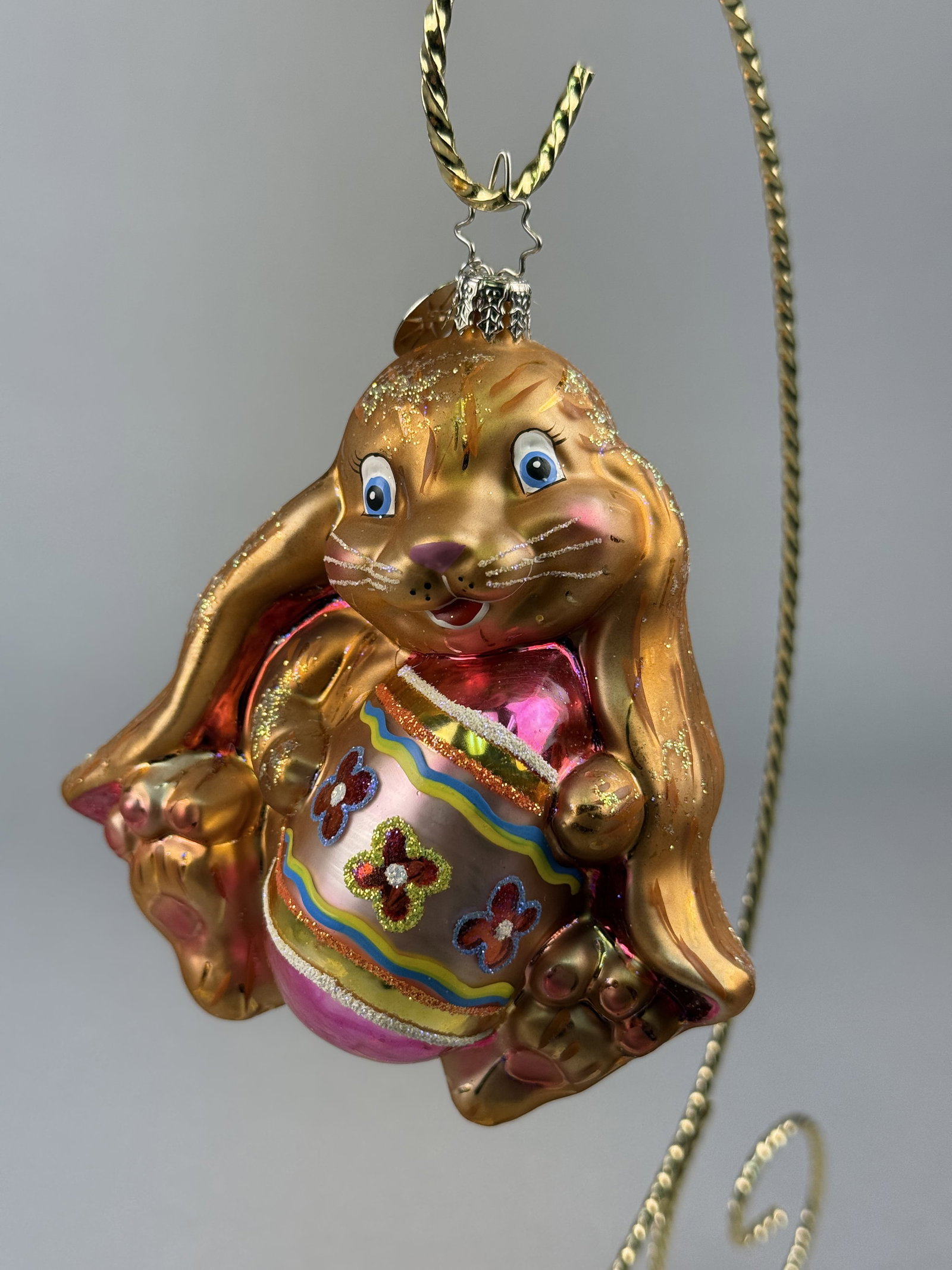 Radko Hoppy Easter Bunny with Decorated Egg Glass Ornament: Stand not included. A hand-blown glass figural ornament produced by Christopher Radko, depicting a seated Easter bunny.The ornament is finished with a decorative metal cap and includes a signature sil