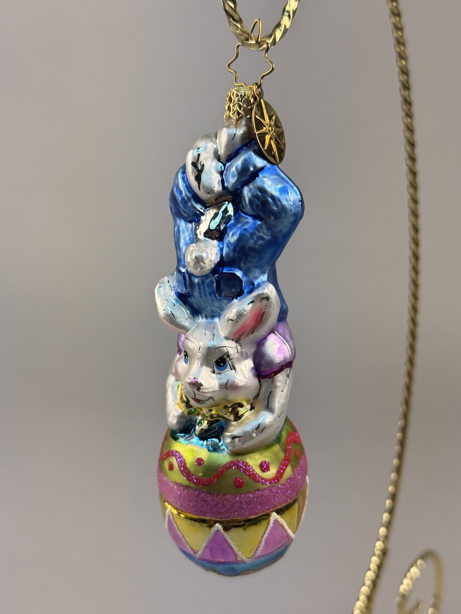 Radko Hop On Top Easter Bunny Glass Ornament: Stand not included. A mouth-blown glass ornament from Christopher Radko titled Hop On Top. It features a gold-toned metal cap and a wire hanger shaped into a six-pointed star. Attached to the hanging 