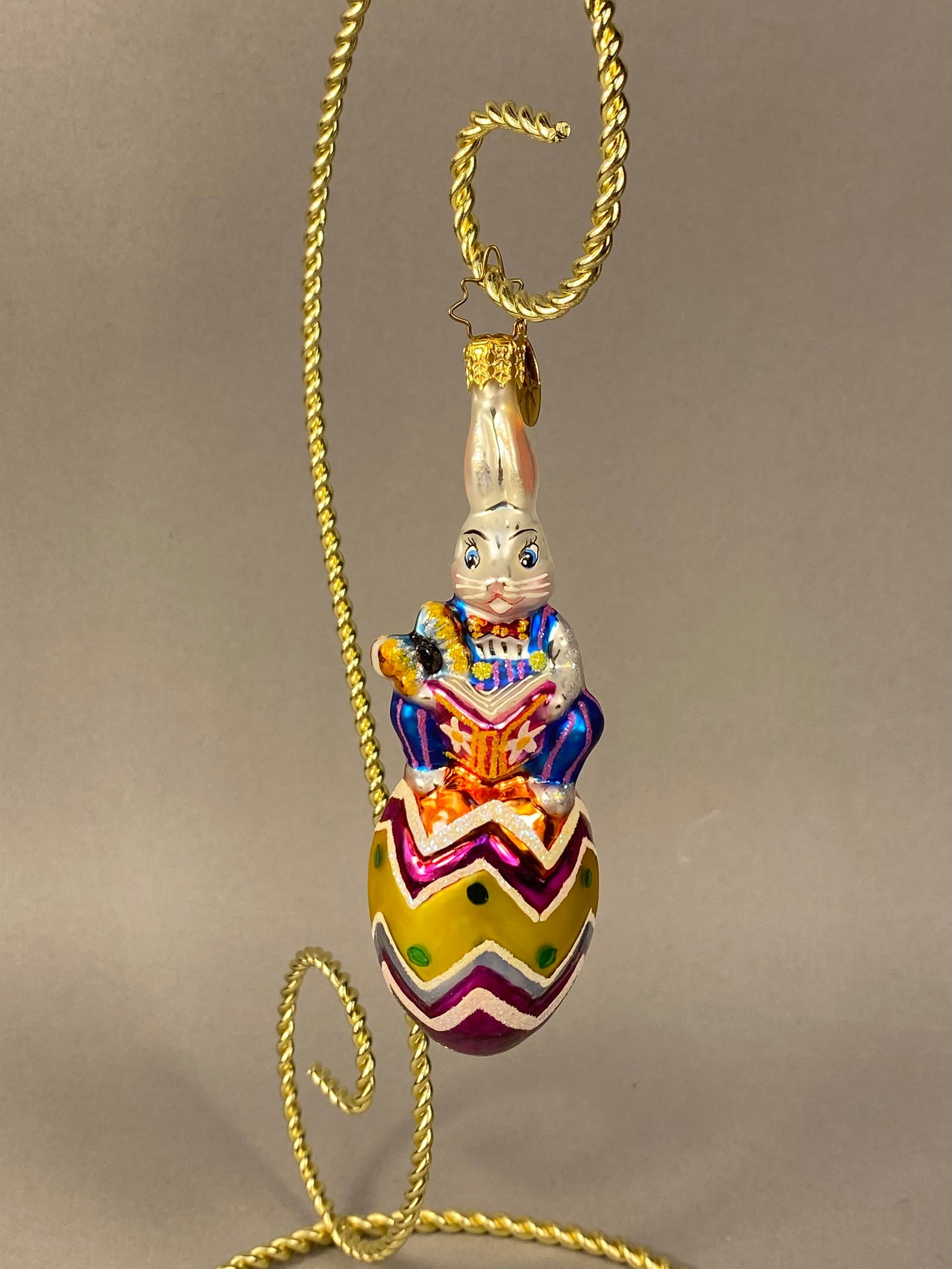 Radko Mask-Hare-Ade Little Gems Glass Rabbit Ornament: Stand Not Included. A hand-blown glass holiday ornament from the Christopher Radko Little Gems collection, titled Mask-Hare-Ade. The piece features a white rabbit wearing blue and pink striped