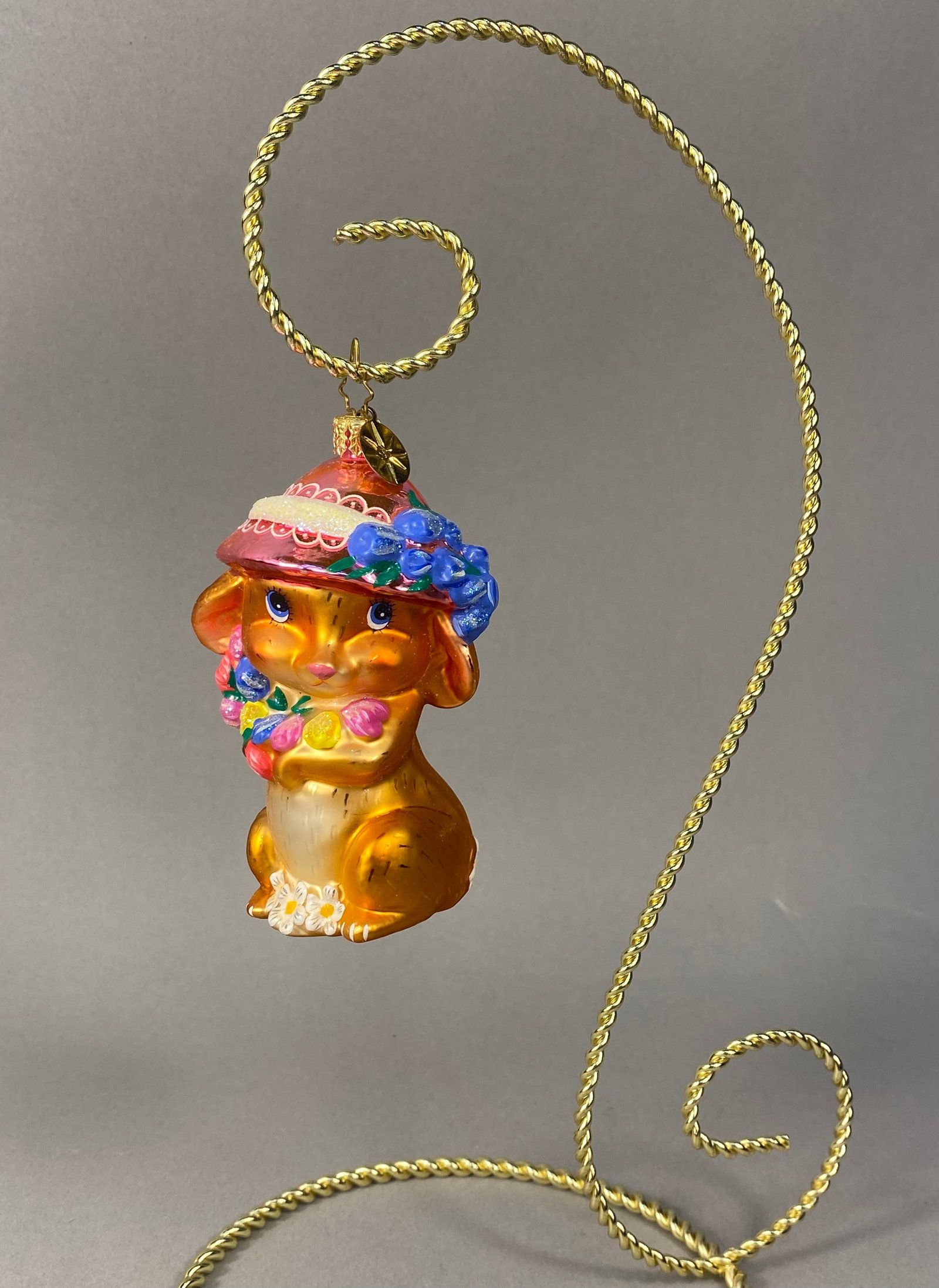 Radko Hand-Painted Blown Glass Rabbit in Floral Bonnet Ornament: Stand Not Included. Christopher Radko hand-painted, mouth-blown glass holiday ornament depicting a stylized rabbit. The figure features an iridescent orange-gold luster finish throughout. The figure i