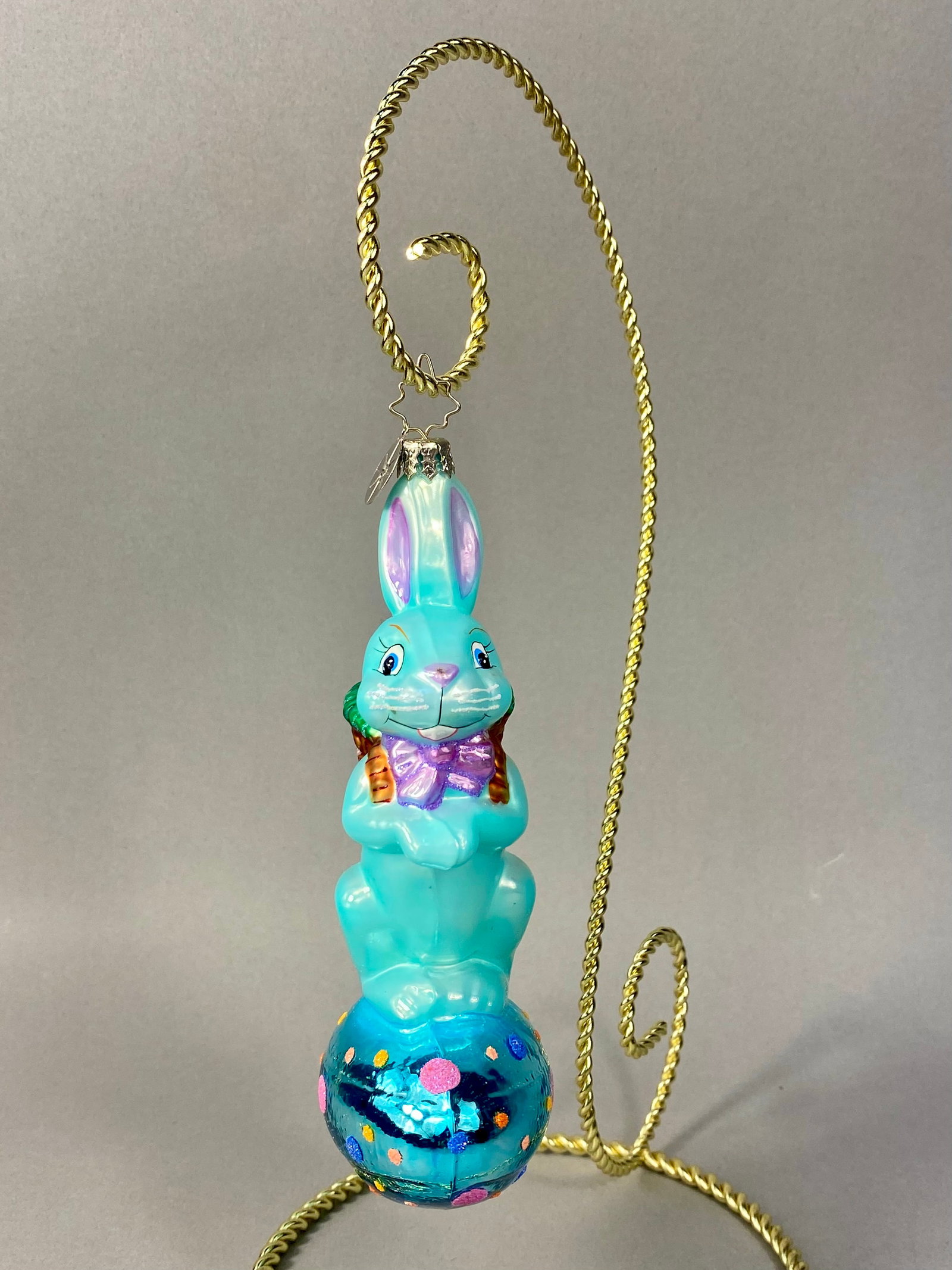 Radko Hand-Painted Glass Easter Bunny on Egg Ornament: Stand Not Included. A mouth-blown and hand-painted glass holiday ornament produced by Christopher Radko. The ornament features a standing light blue rabbit with a pearlescent finish, and carries a