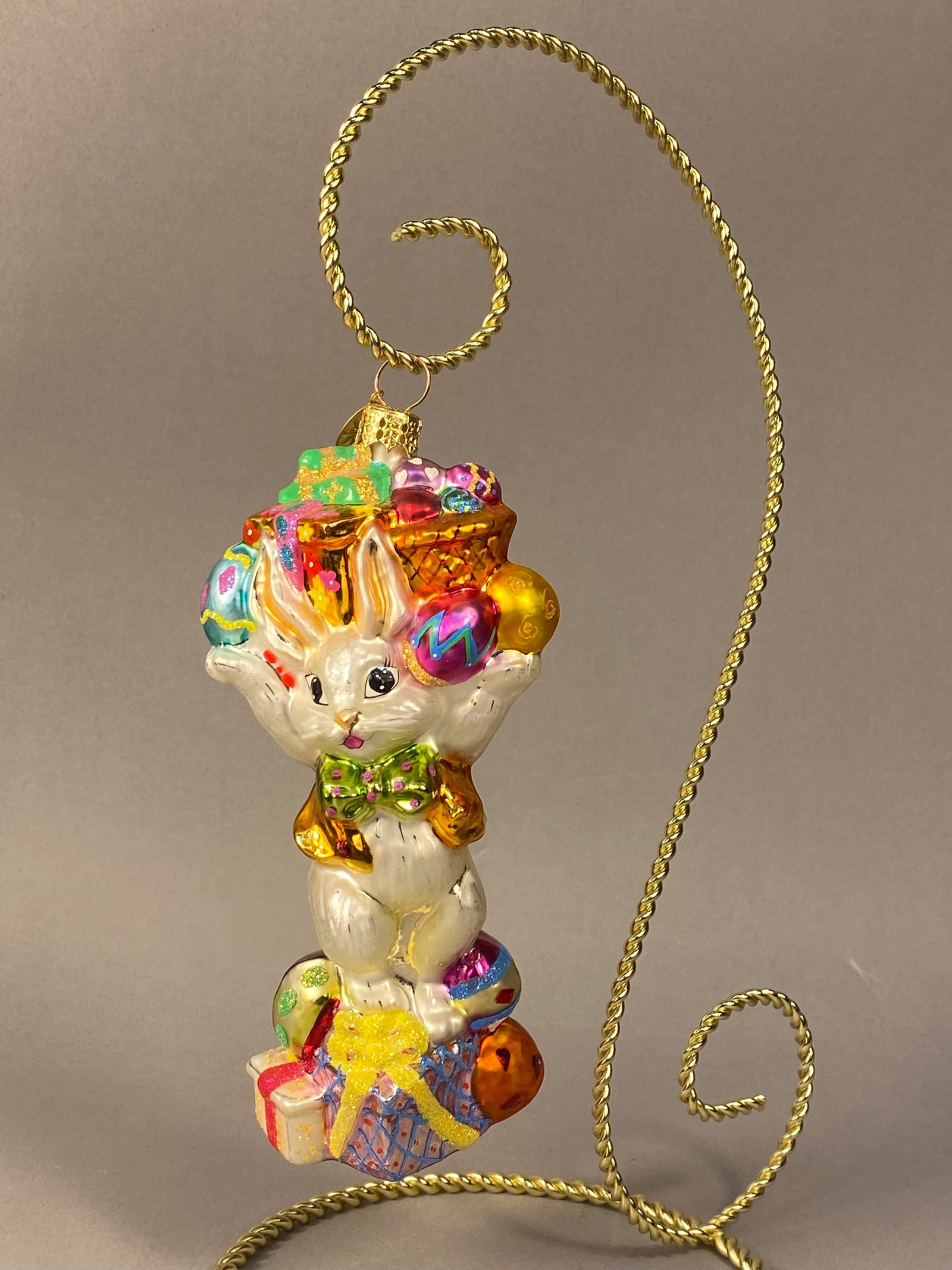 Radko Eggstra Plenty Bunny Glass Ornament: Stand Not Included. A Christopher Radko mouth-blown glass figural ornament titled Eggstra Plenty. The ornament depicts an anthropomorphic white rabbit standing atop a colorful pile of decorated