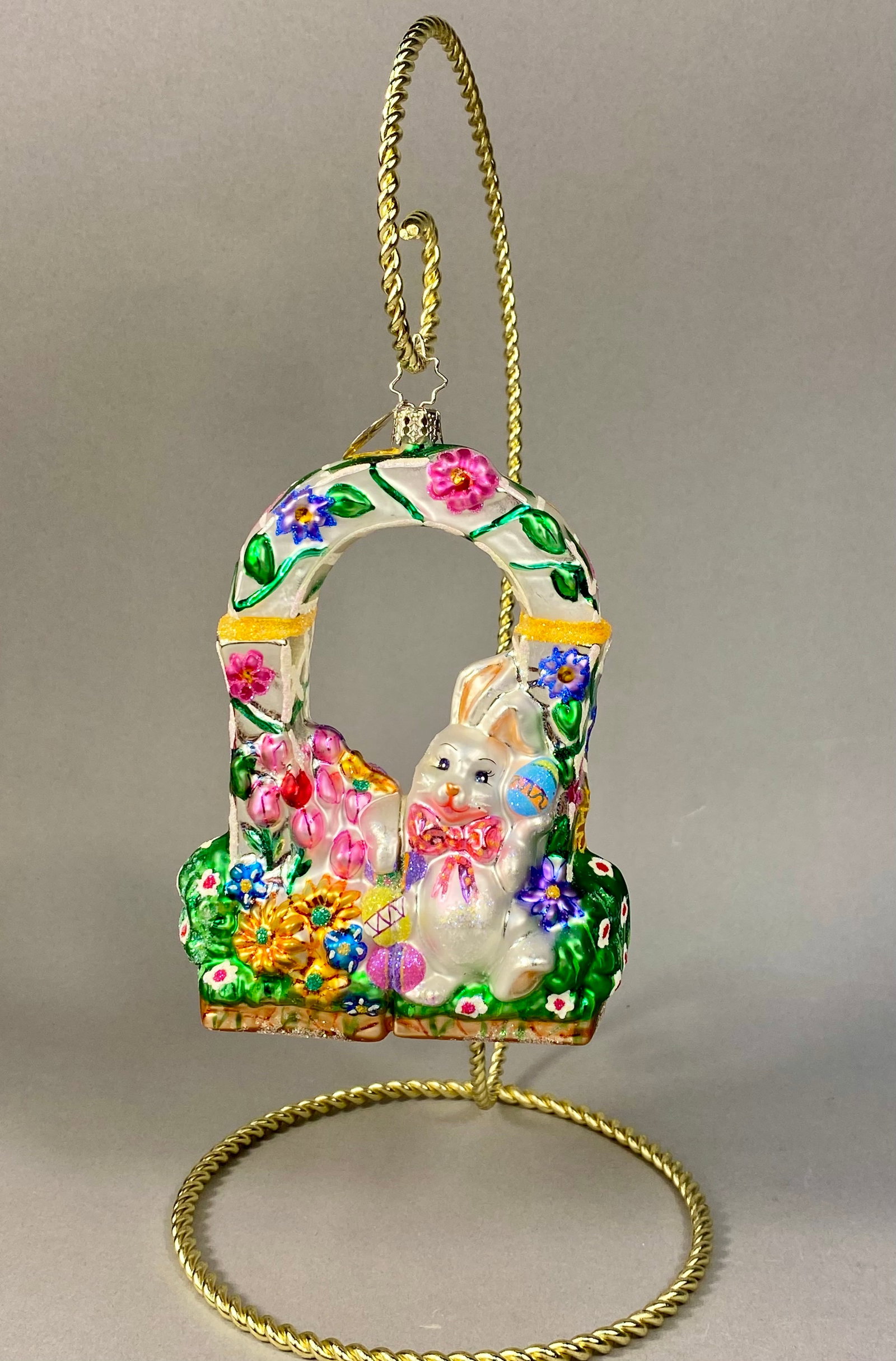 Radko Hoppy Easter Hand-Painted Mouth-Blown Glass Ornament: Stand Not Included. This hand-painted, mouth-blown glass holiday ornament is produced by Christopher Radko and titled Hoppy Easter. The piece features a white rabbit standing within a floral-adorned