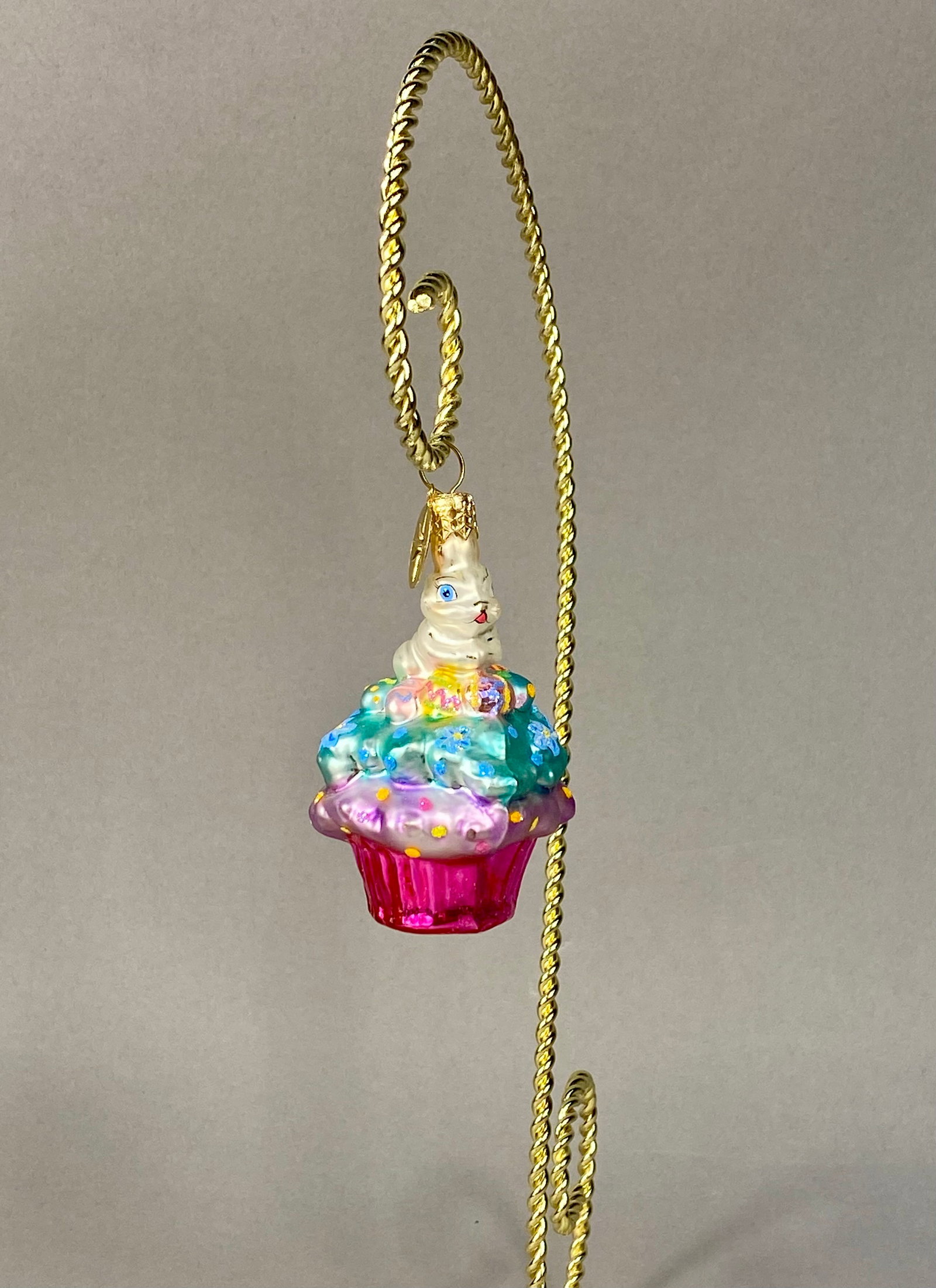 Radko Glass Easter Bunny Cupcake Ornament: Stand Not Included. This is a Christopher Radko hand-painted, mouth-blown glass holiday ornament featuring an Easter theme. The ornament is designed as a cupcake, beginning with a vibrant pink fluted