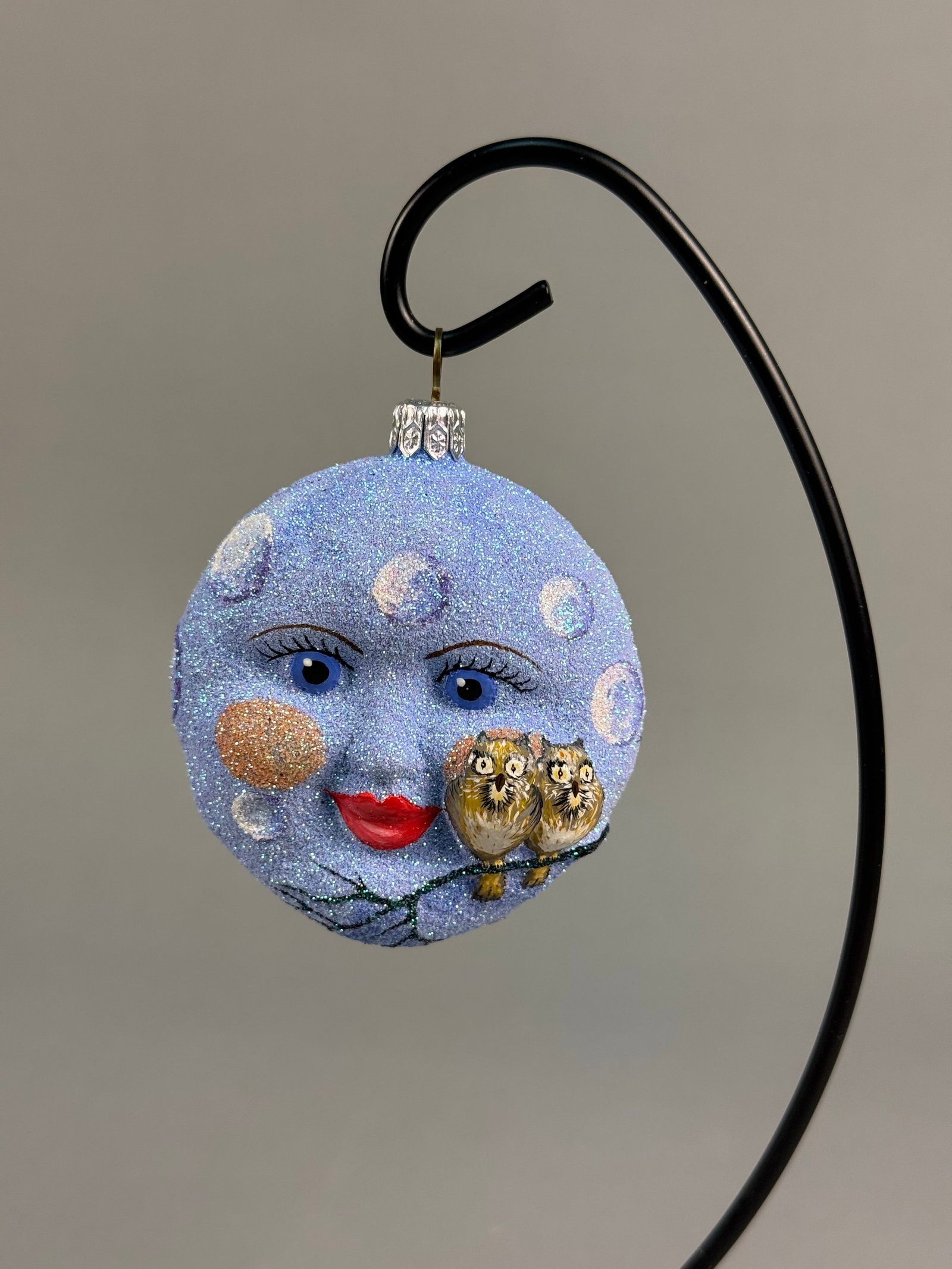 Patricia Breen Moon With Owls Ornament: Stand not included. A hand-decorated light blue glittered Christmas ornament featuring a whimsical moon face with two owls on one side and a bare winter tree on the reverse, measuring approximately 3