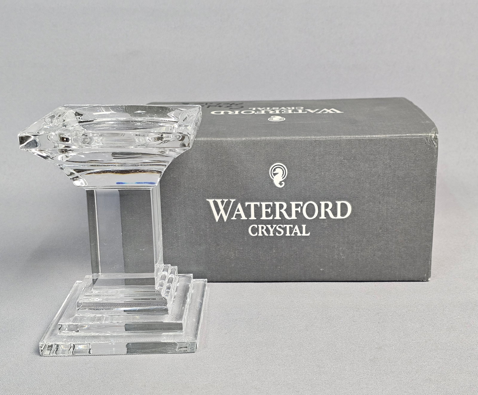 Waterford Crystal Pillar Candle Holder: A lead crystal pillar candle holder manufactured by Waterford in the Metropolitan pattern. This geometric piece features an Art Deco-influenced design with a square tiered stepped base, a solid square