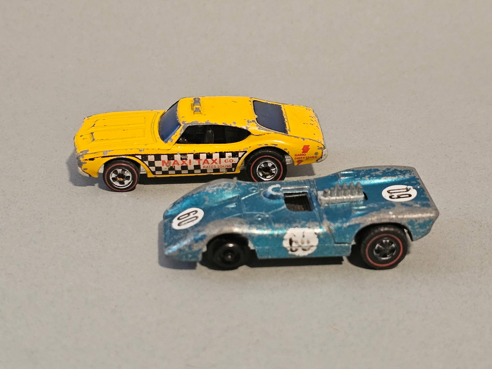 Pair of Vintage Hot Wheels Redline Cars: A pair of vintage die-cast metal toy cars manufactured by Mattel under the Hot Wheels brand during the Redline era. The first car is based on an Oldsmobile 442 casting finished as the Maxi Taxi. It fe