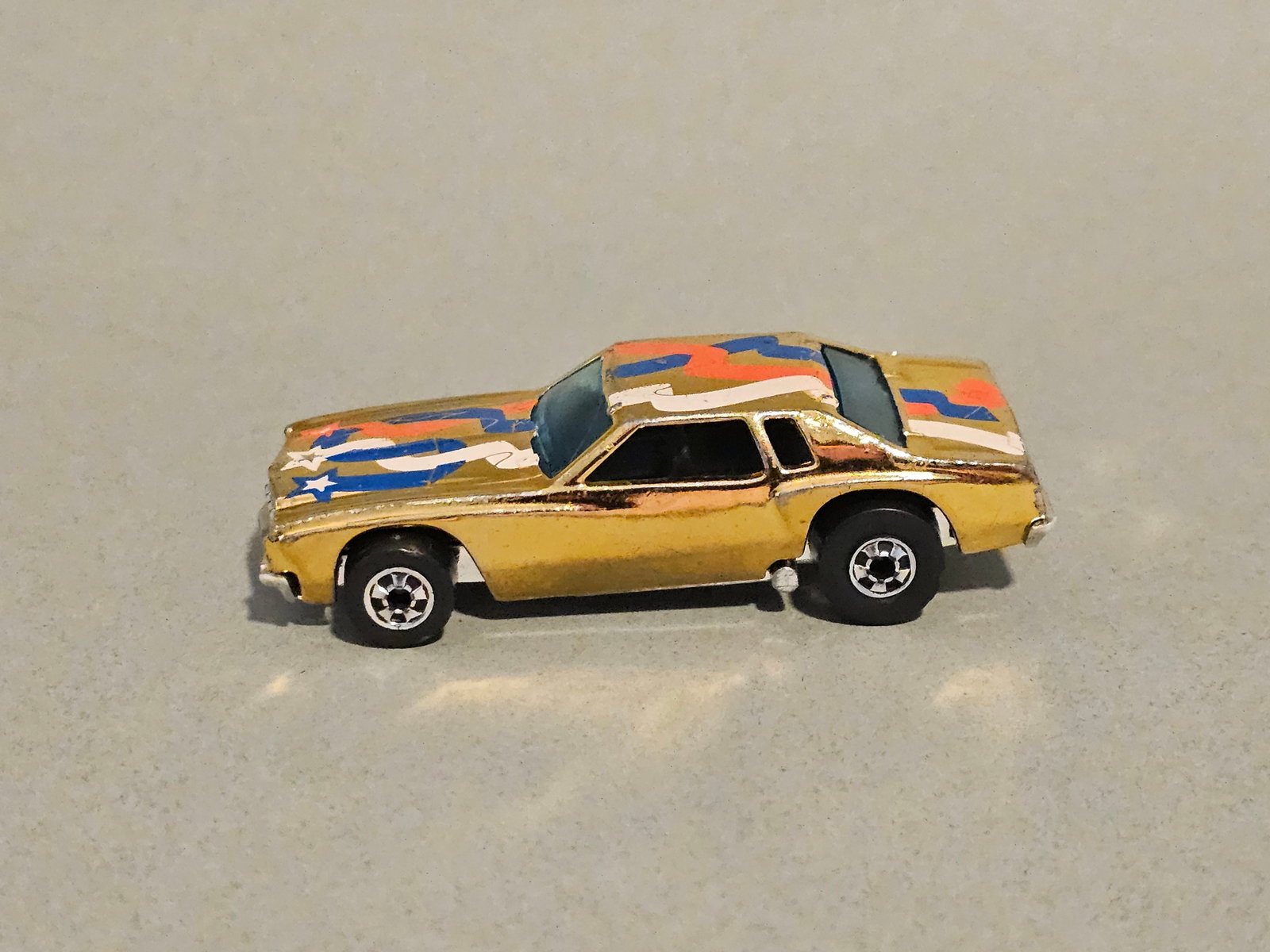 Hot Wheels Lowdown Monte Carlo Car: A vintage Hot Wheels die-cast model car identified as the Lowdown, modeled after a Chevrolet Monte Carlo. This version features a gold chrome-plated finish decorated with a red, white, and blue ribbon