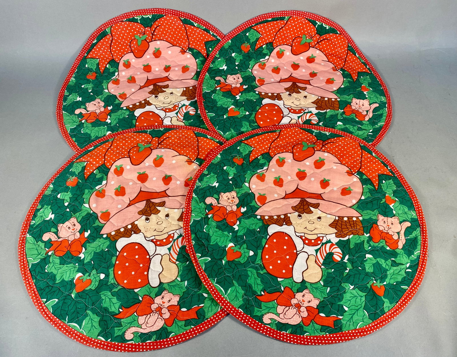 Four Vintage Quilted Strawberry Shortcake Character Placemats: A set of four circular quilted placemats featuring the licensed character Strawberry Shortcake. The central design on each depicts the character in a garden setting, holding a candy cane, accompanied 