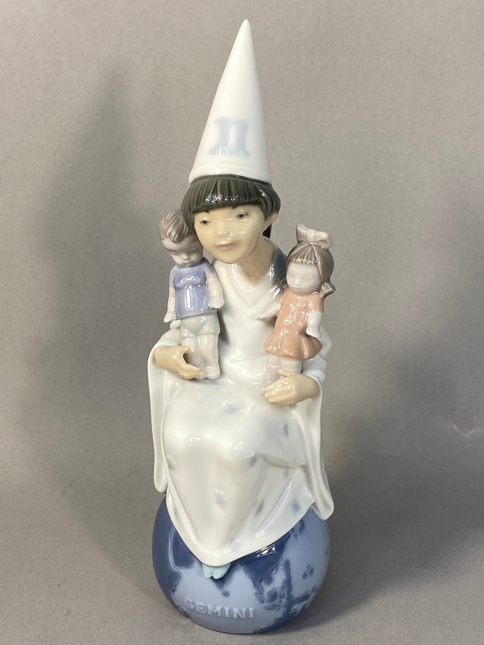 Lladro Porcelain Gemini Zodiac Figurine, Model 6219: A glazed porcelain figurine manufactured by Lladro in Spain, representing the zodiac sign Gemini from the Zodiac Children collection. The figurine depicts a central seated figure wearing a white robe