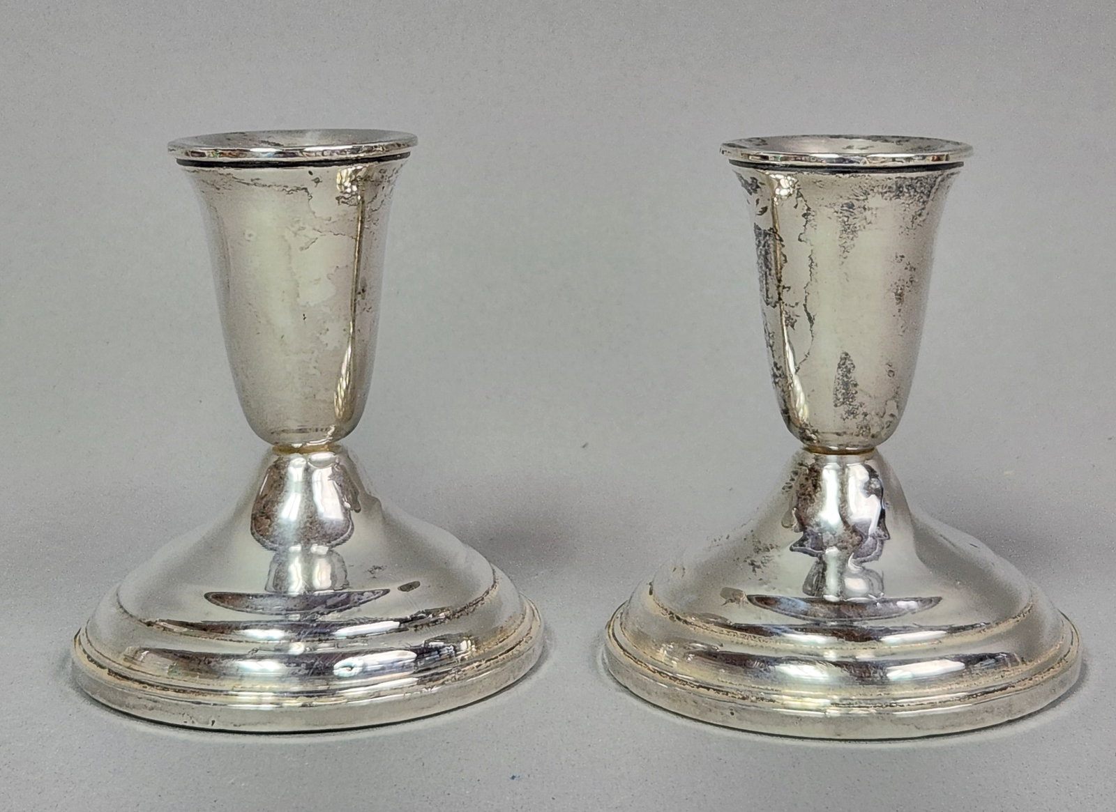 Weighted Sterling Candlesticks: A pair of weighted sterling silver candlesticks in a traditional, low-profile design. Each piece features a tapered candle cup resting on a flared circular pedestal base. The underside of each base be