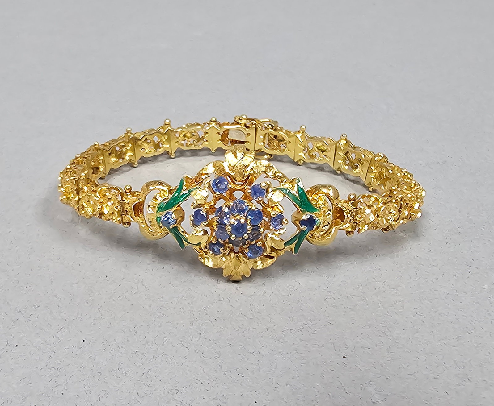 14K Sapphire & Enamel Bracelet 23.5 Grams: The bracelet is 14K yellow gold and the clasp is 18K White Gold. A vintage-style yellow gold bracelet featuring an ornate central openwork motif in a floral and foliate design. The center of the cente
