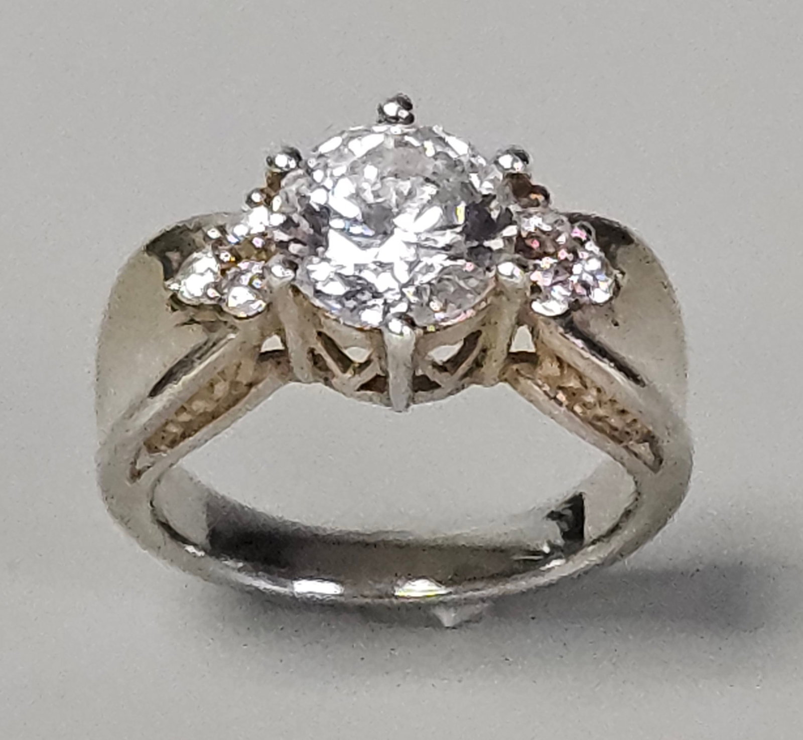 Sterling Colorless Stone Engagement Ring 5.4 Grams: An engagement style ring featuring a central round brilliant-cut colorless stone set in a six-prong setting. The central stone is flanked by six smaller round brilliant-cut colorless stones arranged