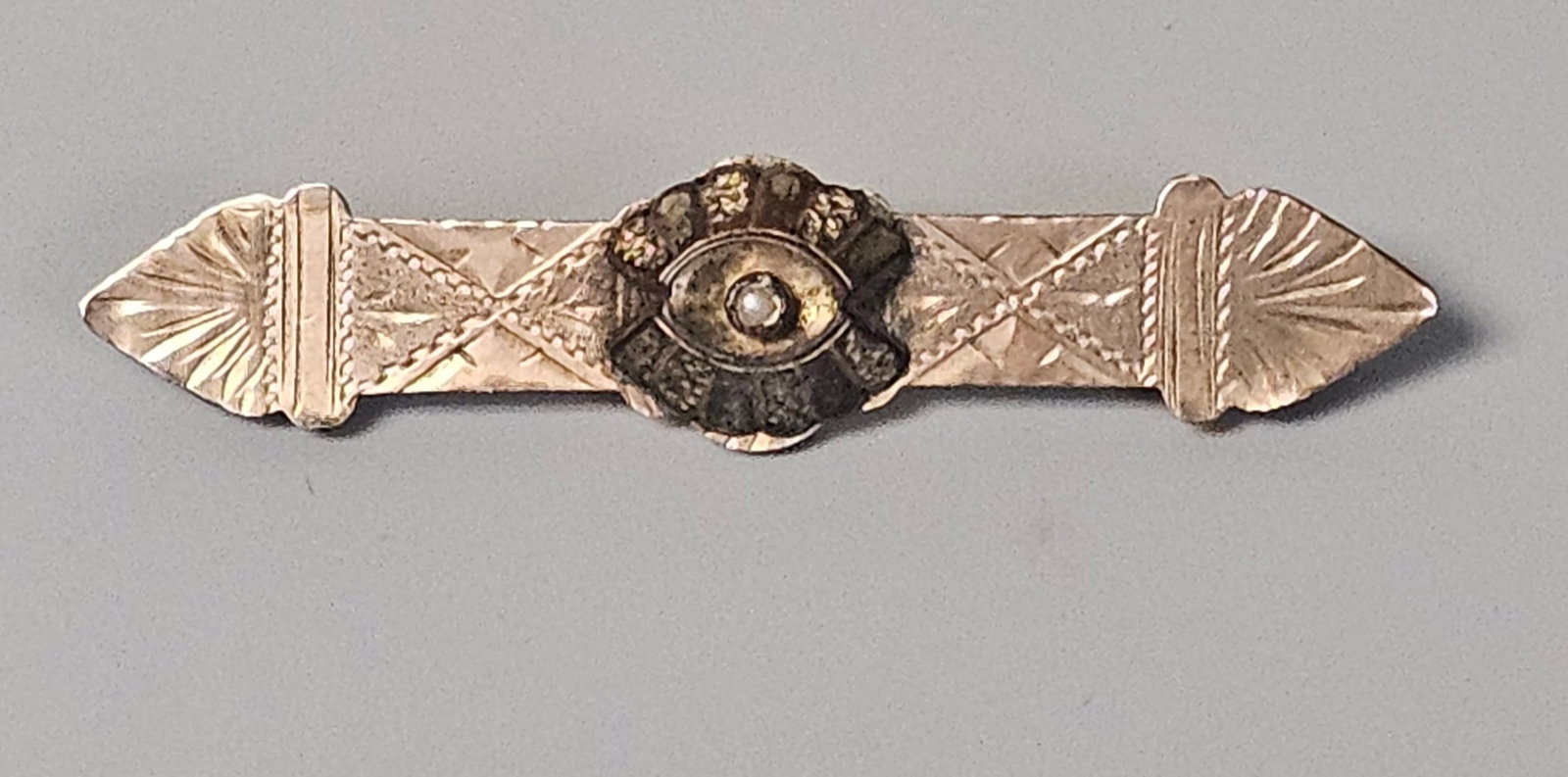 Victorian 12K Gold Filled Bar Brooch Seed Pearl: An antique bar brooch crafted from 12 karat gold-filled metal. The design features a central circular medallion with a raised, textured center set with a single small seed pearl. The tapered bar is de