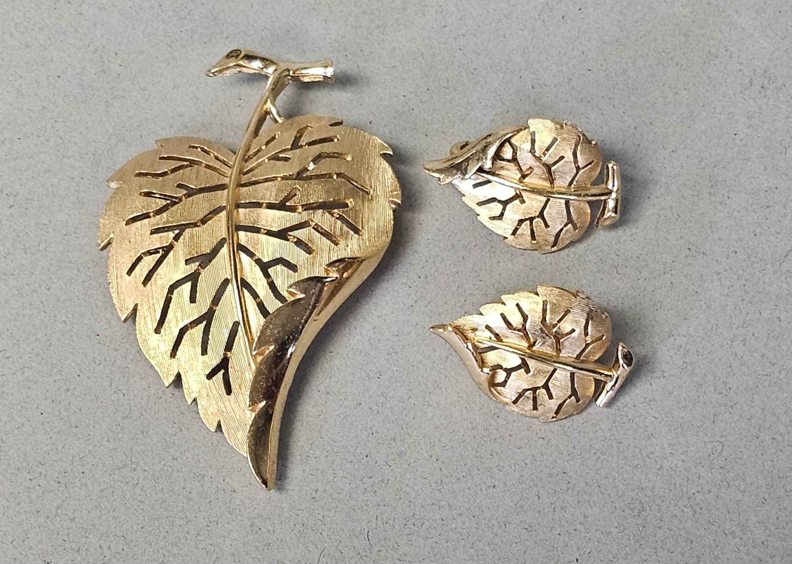 Crown Trifari Mid-Century Brooch & Earrings: A vintage three-piece costume jewelry set by Trifari, consisting of one brooch and a pair of matching clip-on earrings in a botanical leaf motif. The design features a stylized openwork leaf with an i