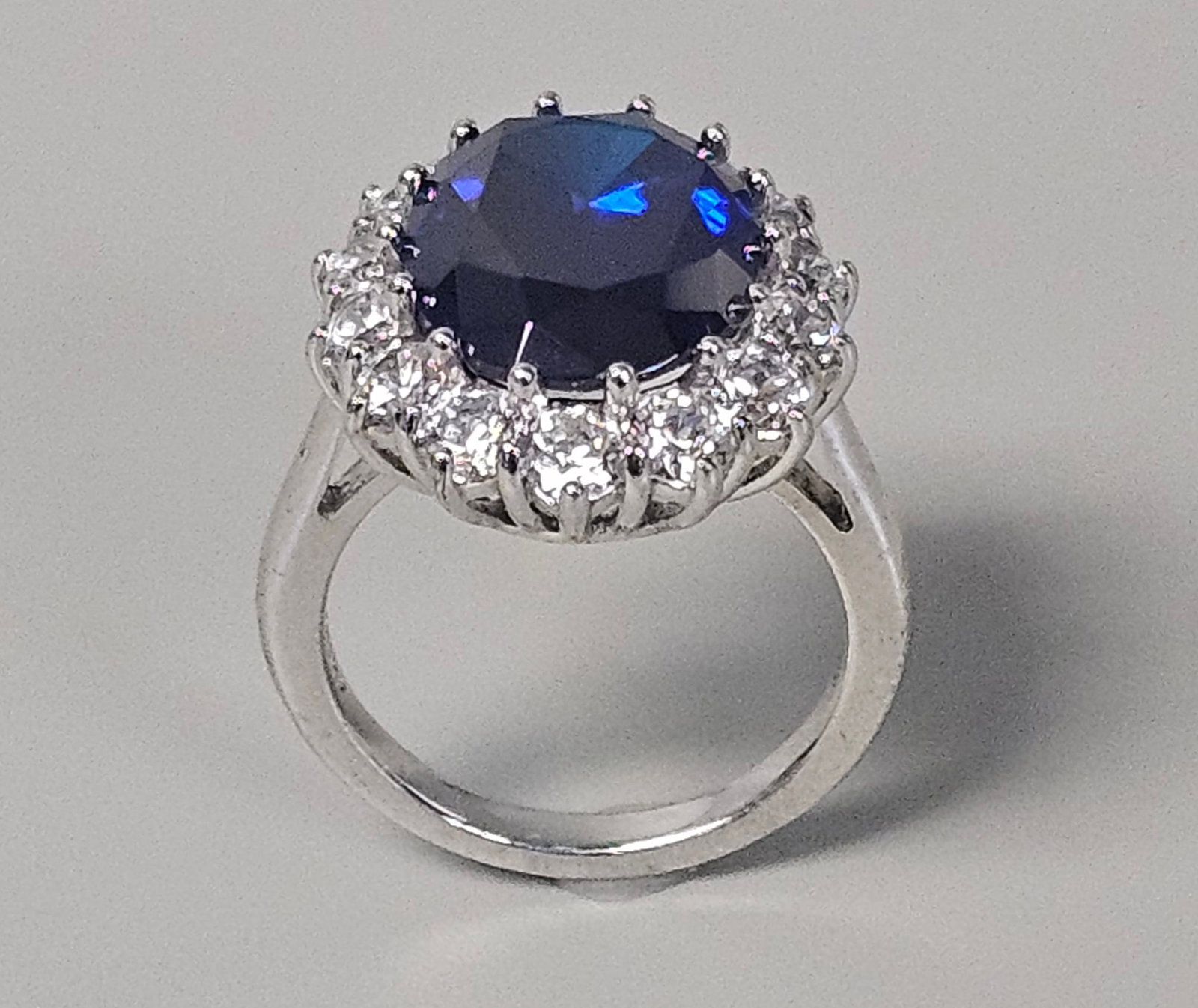 Sterling Cubic Zirconia Oval Halo Ring 7.2 Grams: A sterling silver cocktail ring featuring a large oval-faceted dark blue center stone held in a ten-prong basket setting. The center stone is surrounded by a halo of round-cut clear cubic zirconia sto