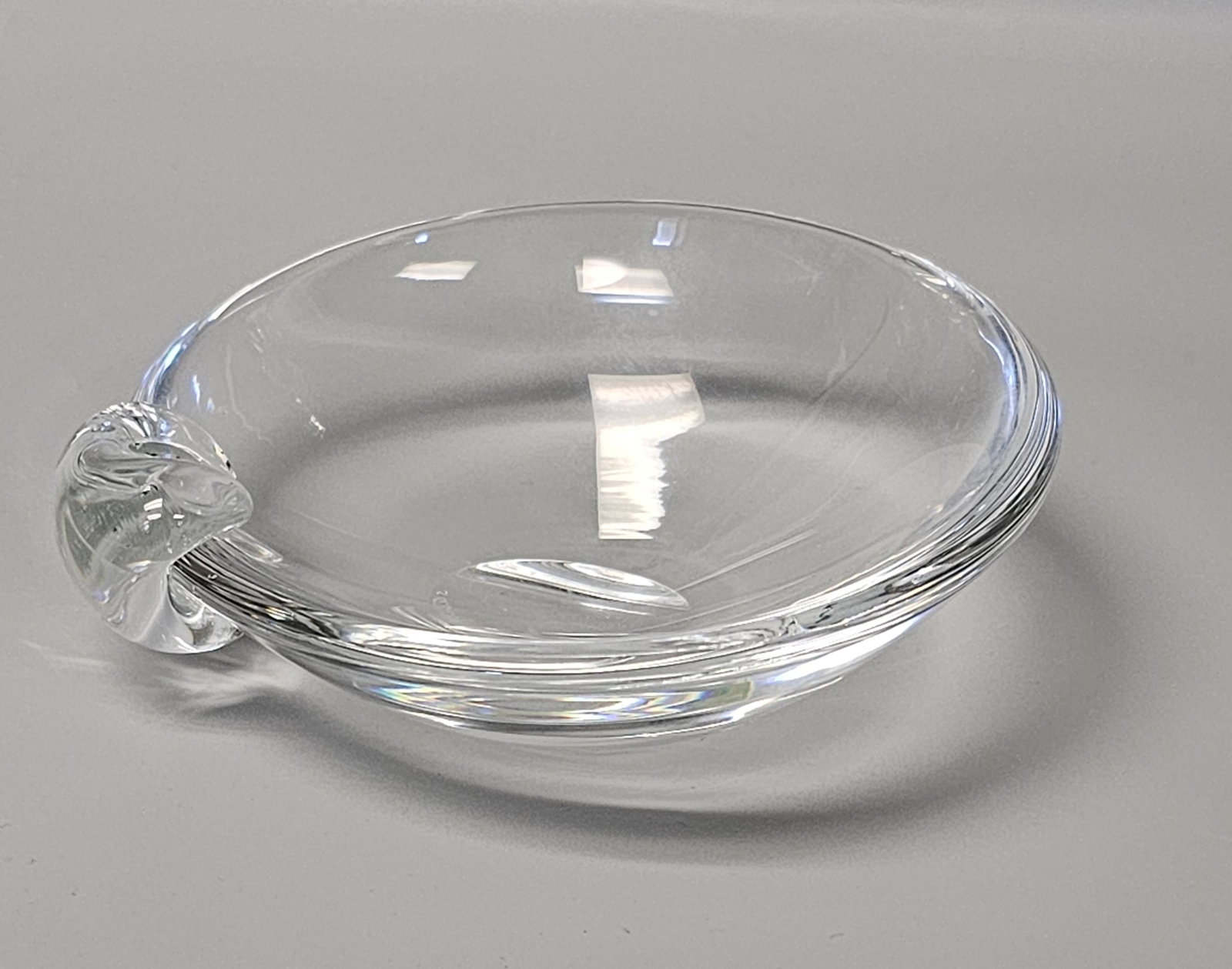 Mid Century Stuben Crystal Ashtray Candy Dish: A hand-blown and hand-formed shallow circular dish crafted from clear crystal by Steuben Glass Works. The vessel features a sloped interior and a thick, rounded rim, accented by a solid glass scrolled