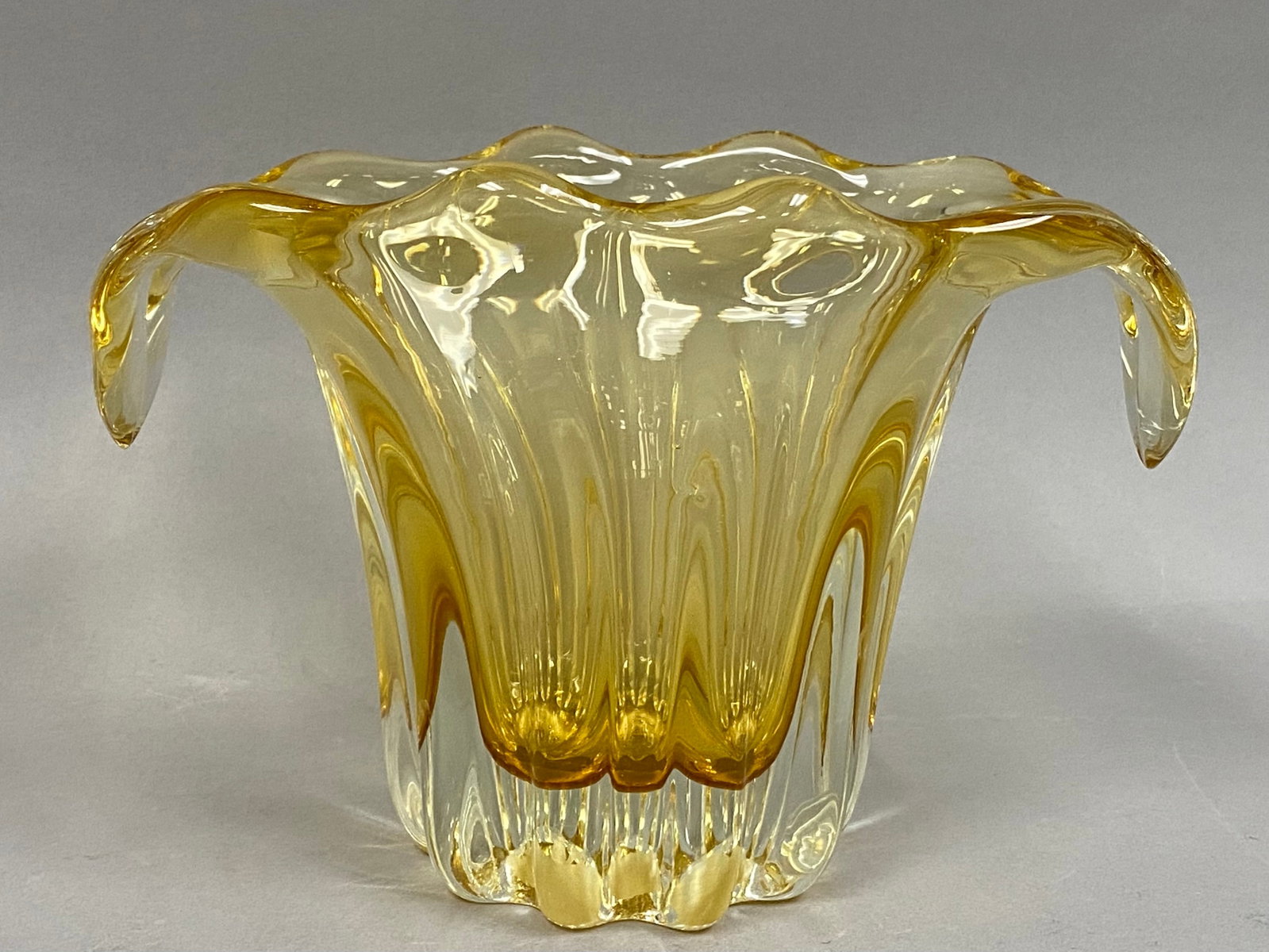 Amber and Clear Glass Vase with Curled Handles: This hand-blown art glass vase features a deeply ribbed body transitioning from a clear, weighted base into a vibrant amber interior. The wide, ruffled rim is accented by two opposing handles that hav