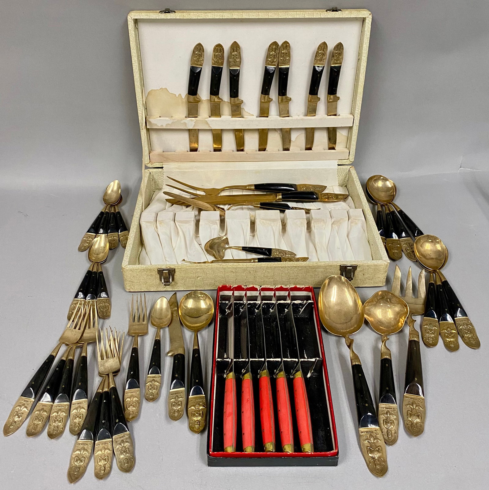 Thai Bronze Flatware & Styson Stainless Steel Knives: A comprehensive flatware service of mid-century Thai export wares and a set of stainless steel steak knives. The Thai service is crafted from polished bronze or brass with dark wood handles secured by