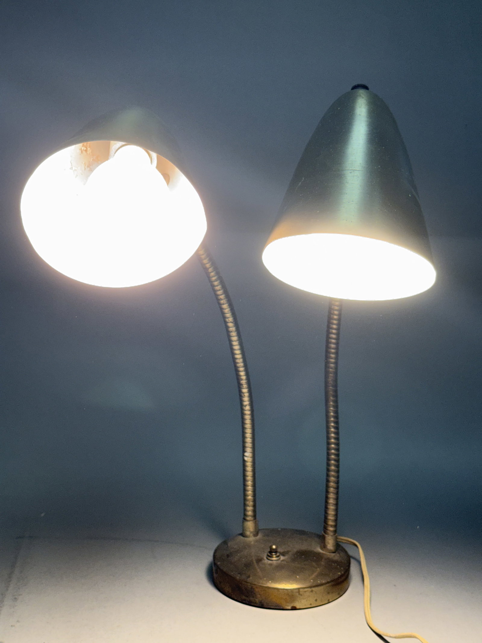 Mid-Century Brass Toned Double Gooseneck Lamp: A vintage Mid-Century Modern double-head gooseneck task lamp, dating from approximately the 1950s to 1960s. The circular metal base is finished to match and includes a central turn-knob switch. The