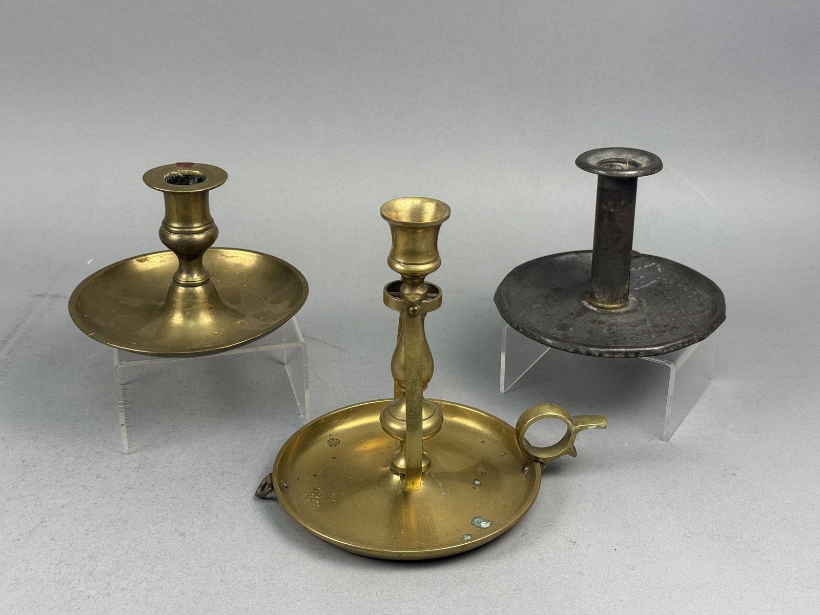 19th Century Style Brass & Pewter Chambersticks: This collection features a trio of antique-style metal chambersticks of varying forms and materials. The third item is a gray-toned metal chamberstick, likely pewter or a similar lead-based alloy,