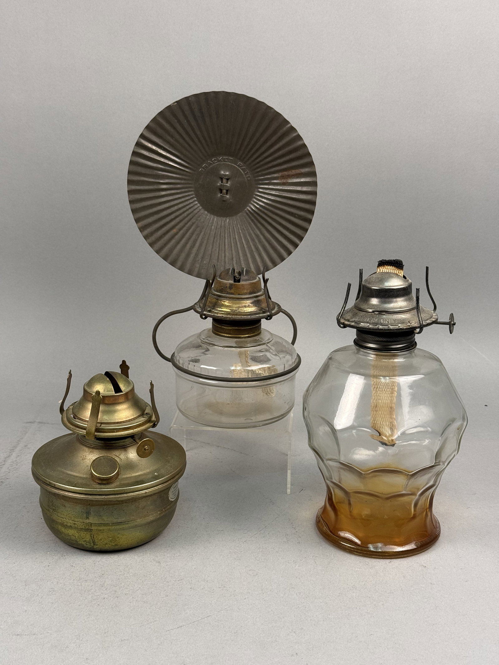 Antique and Vintage Oil & Kerosene Lamps: This lot comprises a collection of three non-electric combustion lamps of varying designs. Included is a bracket-style hand lamp featuring a bulbous clear glass reservoir held within a wire frame,