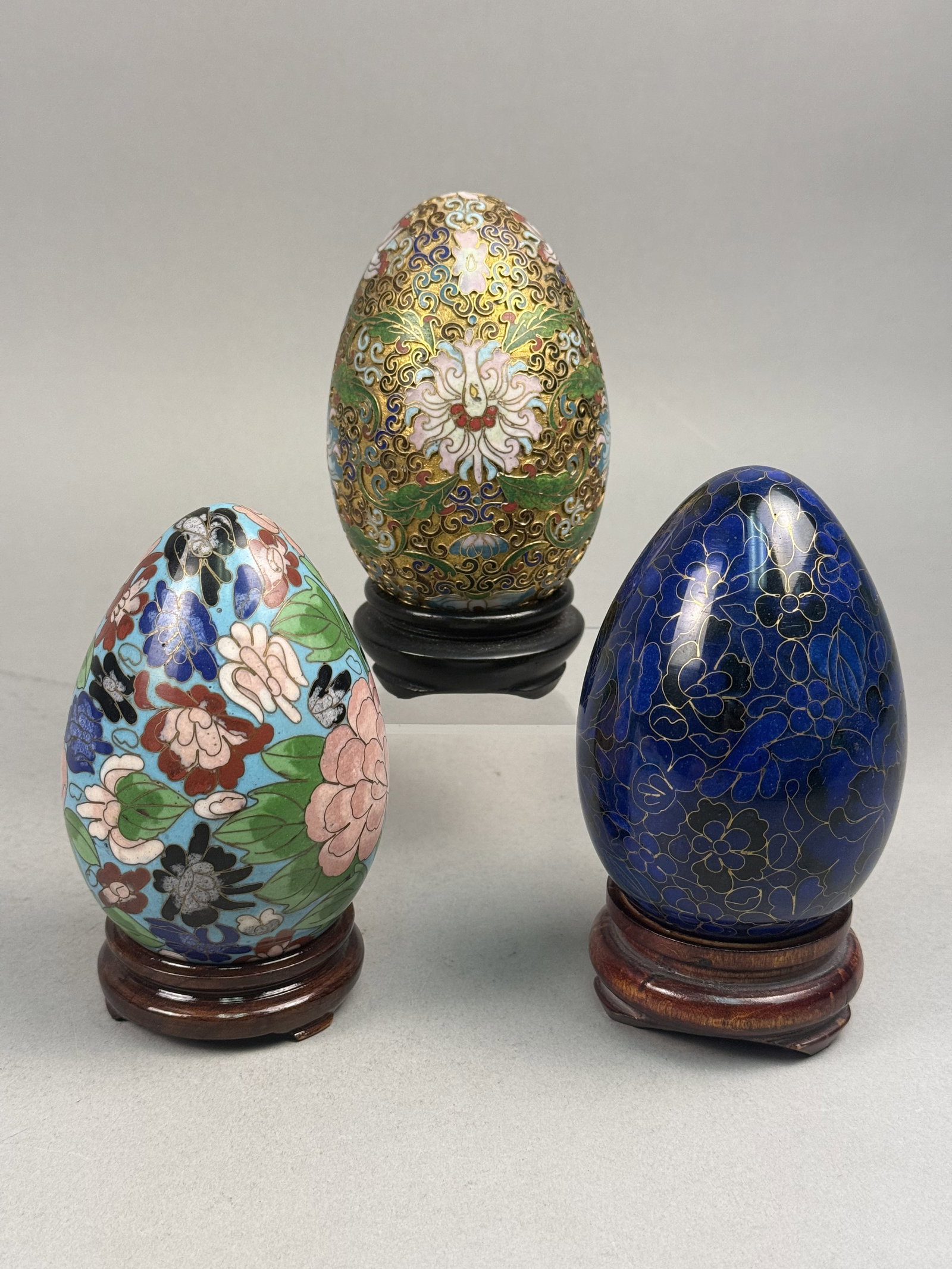 3 Chinese Cloisonne Enamel Decorative Eggs: A trio of decorative eggs featuring Chinese cloisonne enamel work in three distinct styles. Each egg is constructed using the traditional cloisonne technique, where thin metal wires are soldered to a