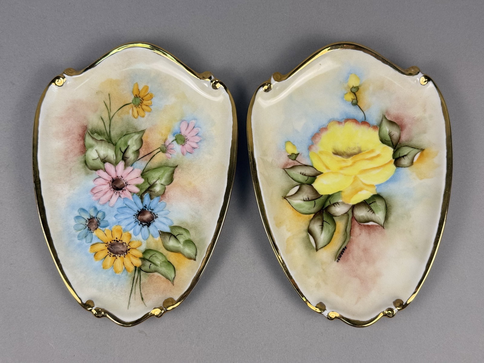 Hand-Painted Porcelain Wall Plaques A. Grambo: This is a pair of porcelain wall plaques featuring shield-shaped forms with scalloped edges and gold-gilt borders. The plaque with the rose is signed A. GRAMBO in black script on the front near the