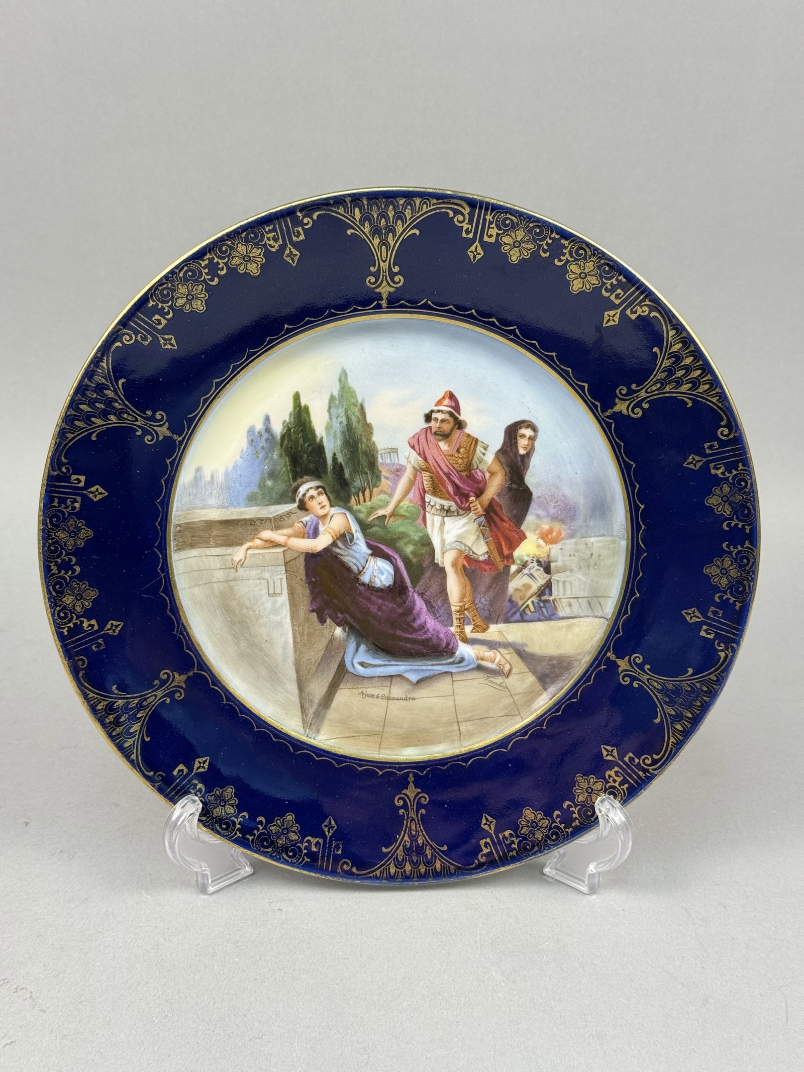 Royal Vienna Style Porcelain Plate: This decorative porcelain cabinet plate features a hand painted central vignette and an ornate gilded border. The wide cobalt blue rim is embellished with intricate gold scrolling foliate patterns,