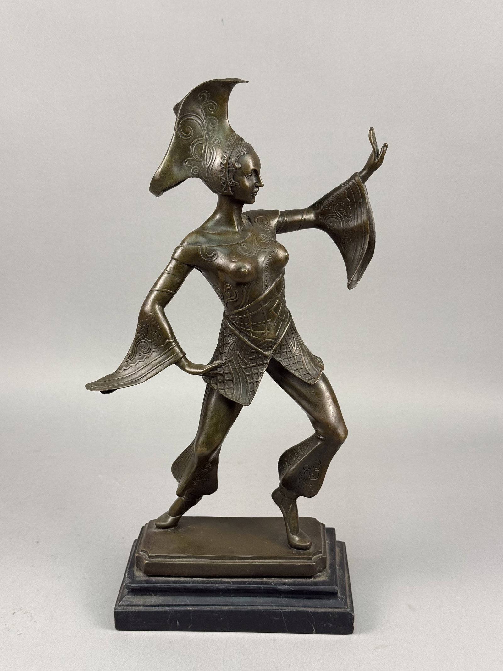 Art Deco Style Bronze Dancer Sculpture: The bronze is finished with a variegated dark brown and olive green patina. The figure stands on an integral stepped bronze base, which is mounted atop a two-tiered black marble plinth. The underside